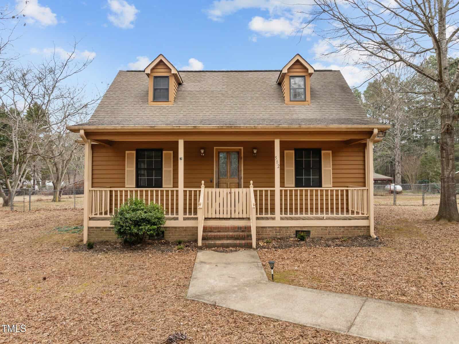 Nestled at 582 Pollyanna Road in Henderson, a brown wooden house with two dormer windows and a cozy front porch is embraced by leafless trees. The porch features a railing and steps descending to a concrete pathway, under a clear sky dotted with clouds.