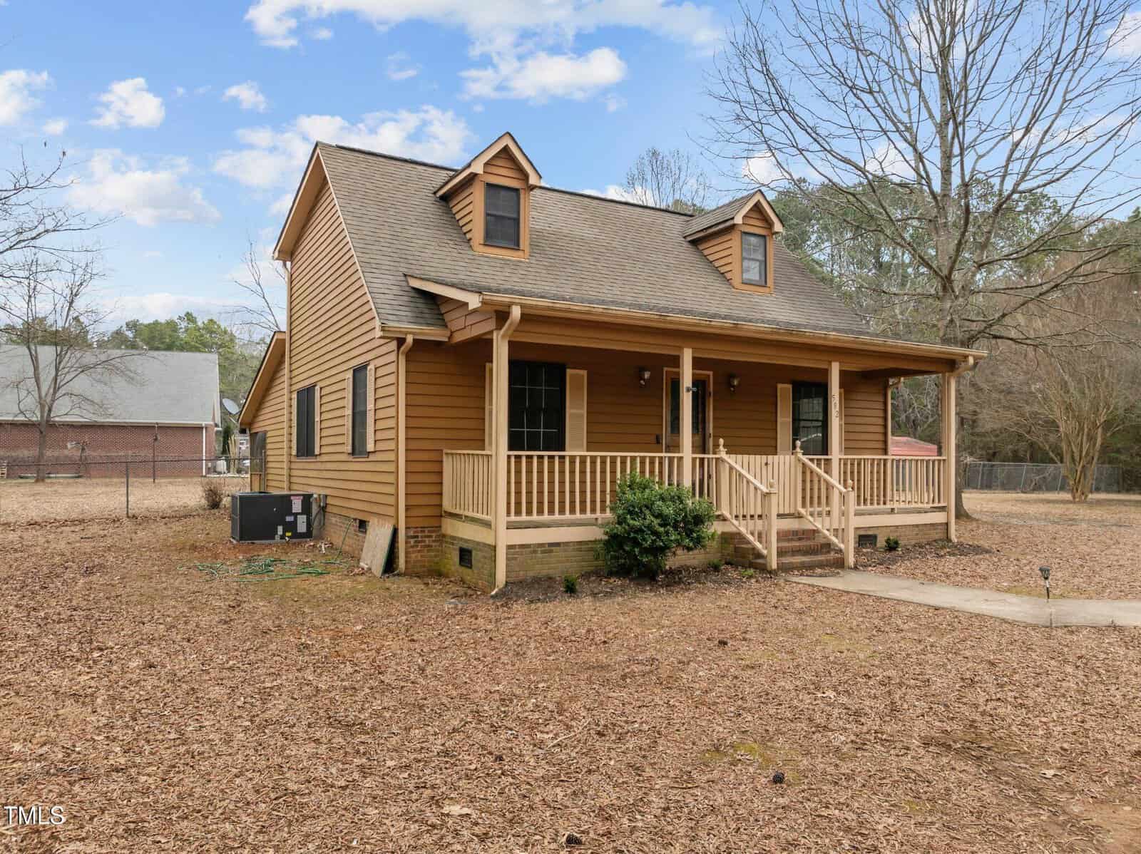 A two-story house with a wooden exterior and a covered front porch. The roof has two dormer windows, and there's a small set of stairs leading to the entrance. The yard is covered in dry leaves, with a leafless tree nearby.