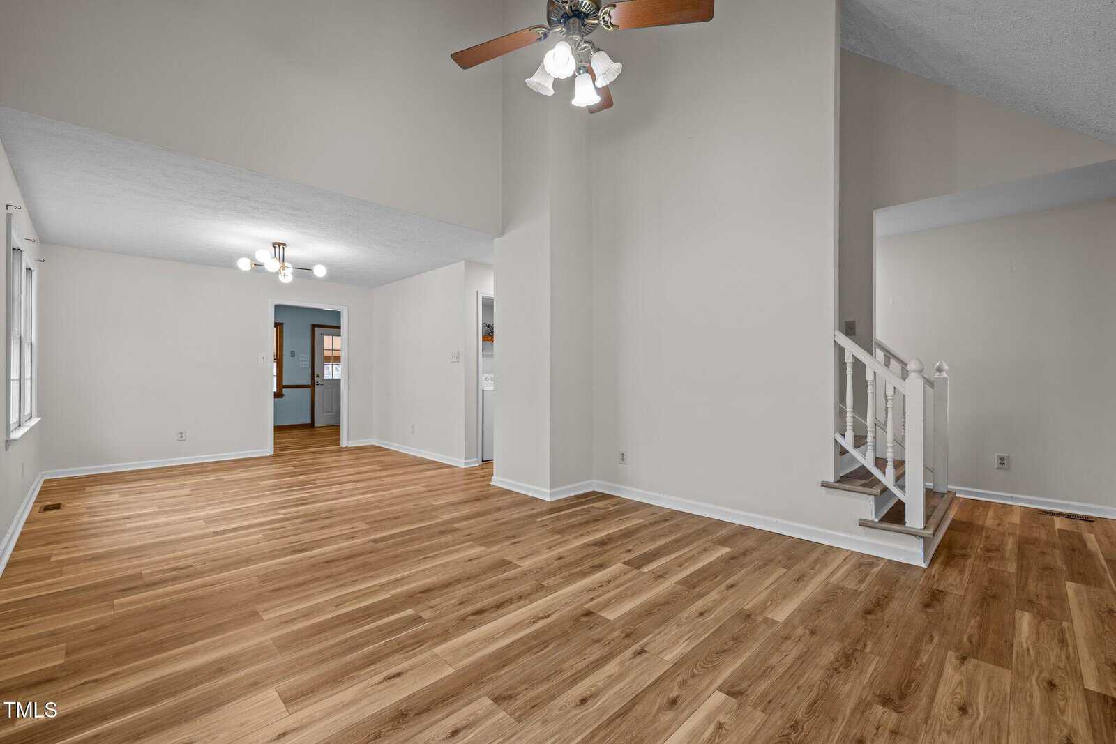 Spacious empty room with wooden flooring and light beige walls. Ceiling fans hang from the ceiling; stairs with white banisters lead to an upper level. Large windows allow natural light, and a doorway connects to another room in the background.