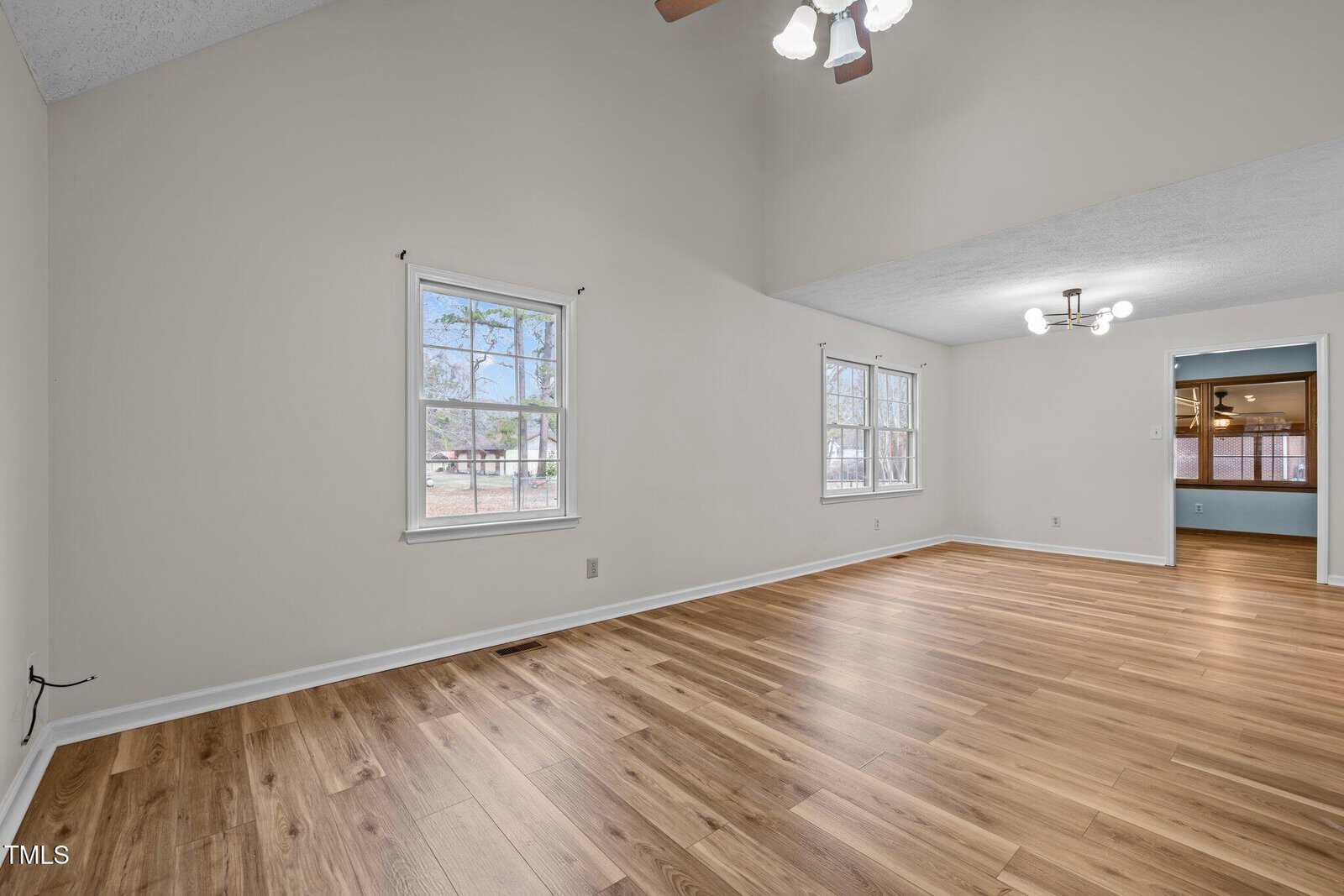 A spacious, unfurnished room with light wood flooring and cream walls. Two ceiling fixtures provide lighting. Two windows on the left wall let in natural light. An open doorway leads to another room with darker flooring.