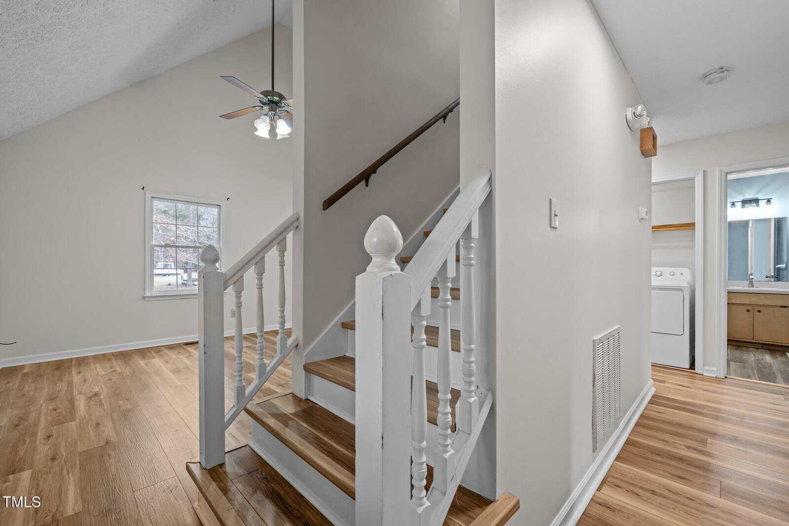 A small staircase with white balusters and wooden steps leads to an upper level. The foreground shows a room with wood flooring. A ceiling fan is visible above. In the background, there is a laundry area with a washing machine and a bathroom.