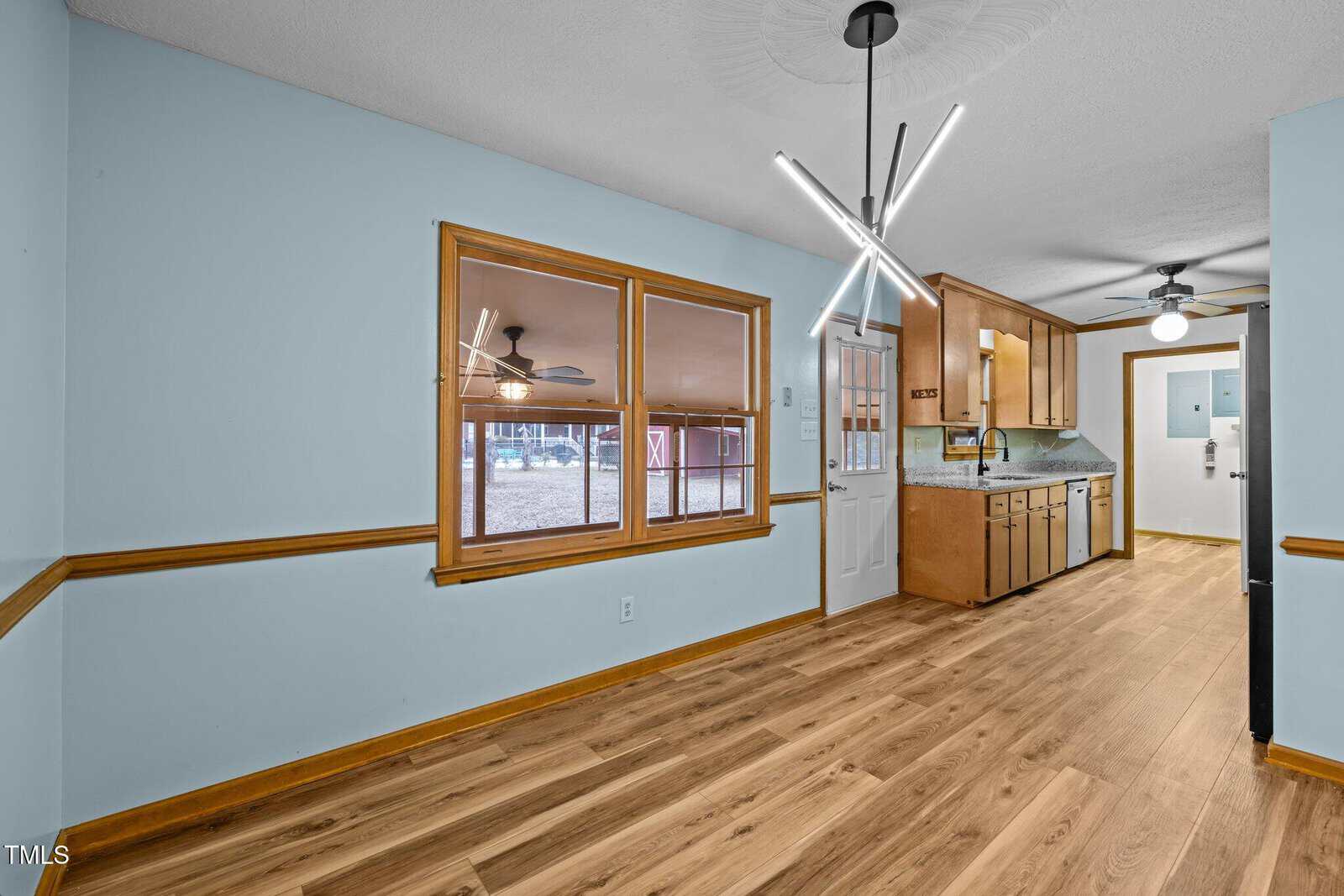 A room with light blue walls and wood flooring features a modern light fixture. There are wooden cabinets and a white door with a window. Outside the window, a red barn is partially visible. Ceiling fans are seen in both the kitchen and adjacent room.