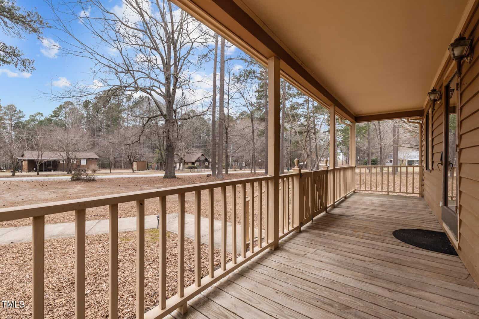 A wooden porch with railings extends along the side of a house, overlooking a yard with bare trees. A pathway leads to other wooden structures in the background under a partly cloudy sky.