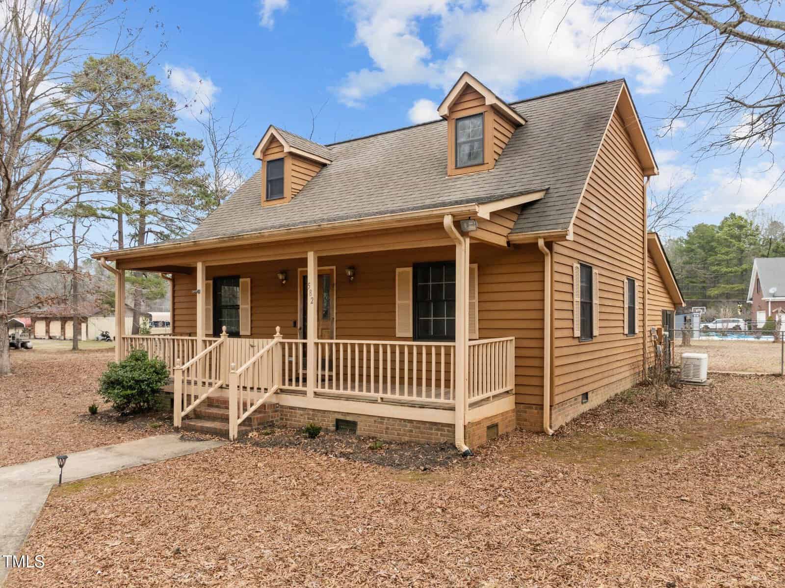 A small, brown wooden house with a porch and gabled roof is surrounded by bare trees and a grassy yard. There are two dormer windows on the roof and several steps leading up to the porch, with a sidewalk extending to the left.