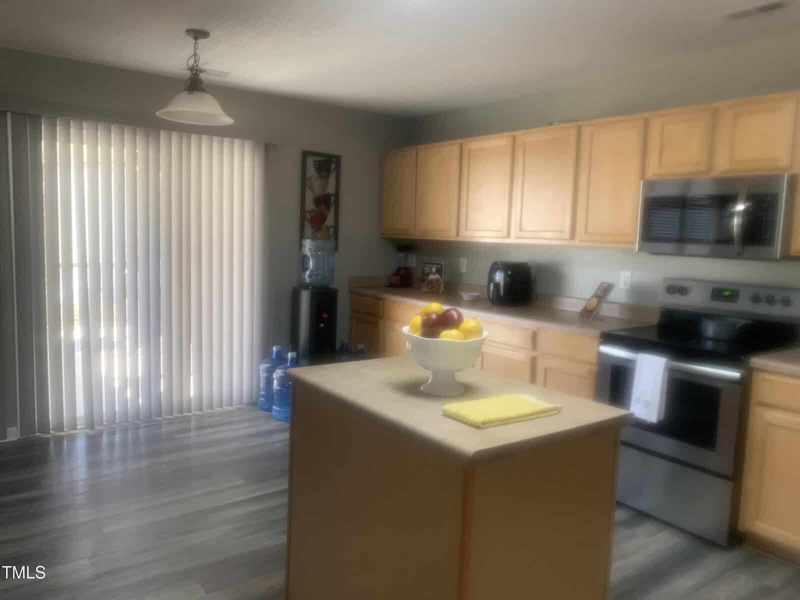 Kitchen with wooden cabinets, stainless steel appliances, and a central island. A bowl of fruit and a yellow towel are on the island. Vertical blinds cover the sliding door on the left. A water cooler and bottles are beside the door.
