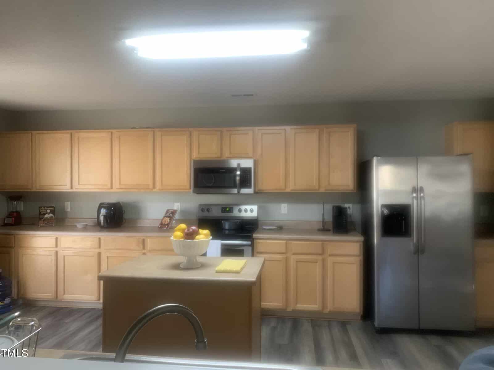 A kitchen with wooden cabinets, a stainless steel refrigerator, and a built-in microwave above the stove. An island counter features a bowl of fruit and a yellow cloth. The floor is wood, and overhead lighting is on.