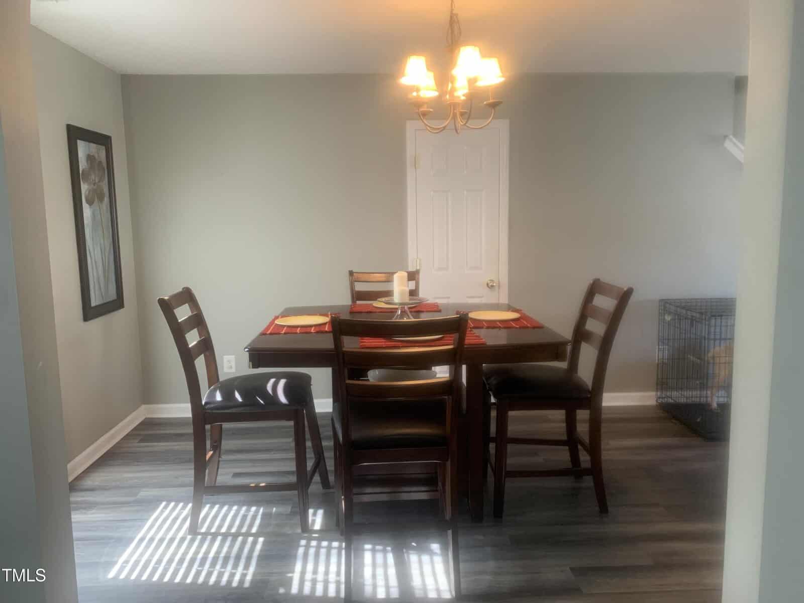 A dining room with a square wooden table set for four, featuring brown chairs and red placemats. A chandelier hangs above. The floor is dark wood, and there's a large framed artwork on the left wall. A dog crate is visible in the right corner.