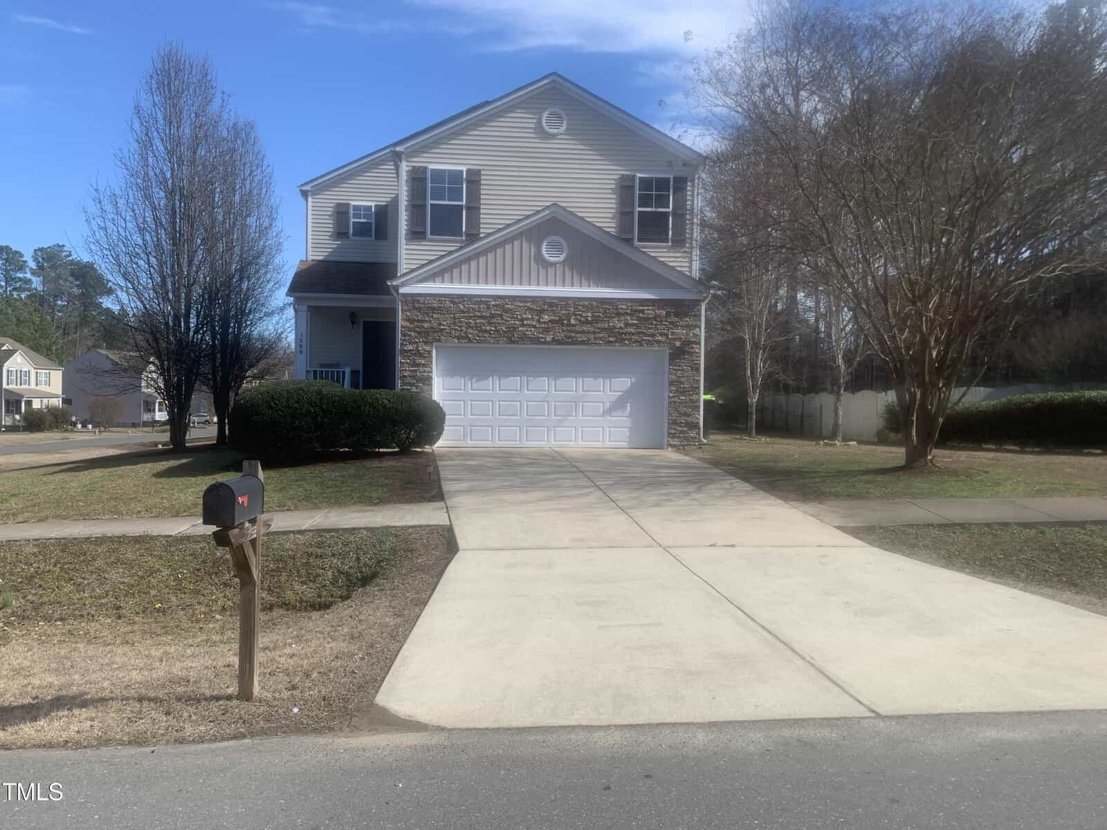 Located at 1208 Shining Water Lane in Creedmoor, this charming two-story house with beige siding and stone accents features a white garage door and a concrete driveway. Leafless trees frame the mailbox on the grassy front lawn, all under a clear blue sky.