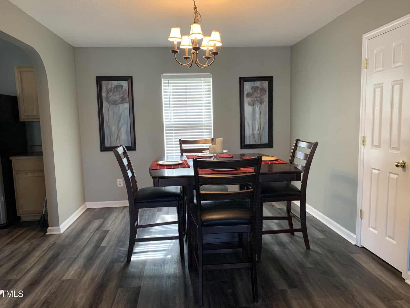 A dining room features a wooden table with four matching chairs. Above the table hangs a chandelier. The room has two large framed floral artworks on the walls and a window with blinds partially open. The floor is dark wood, and there's a closed white door.