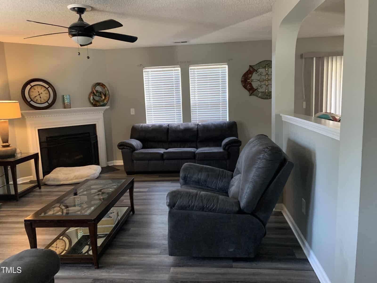 A living room with gray walls, featuring a black leather sofa and recliner. A glass coffee table sits on a wooden floor. A fireplace with a clock on the mantel is in the corner. Two windows with blinds are behind the sofa. A ceiling fan hangs above.