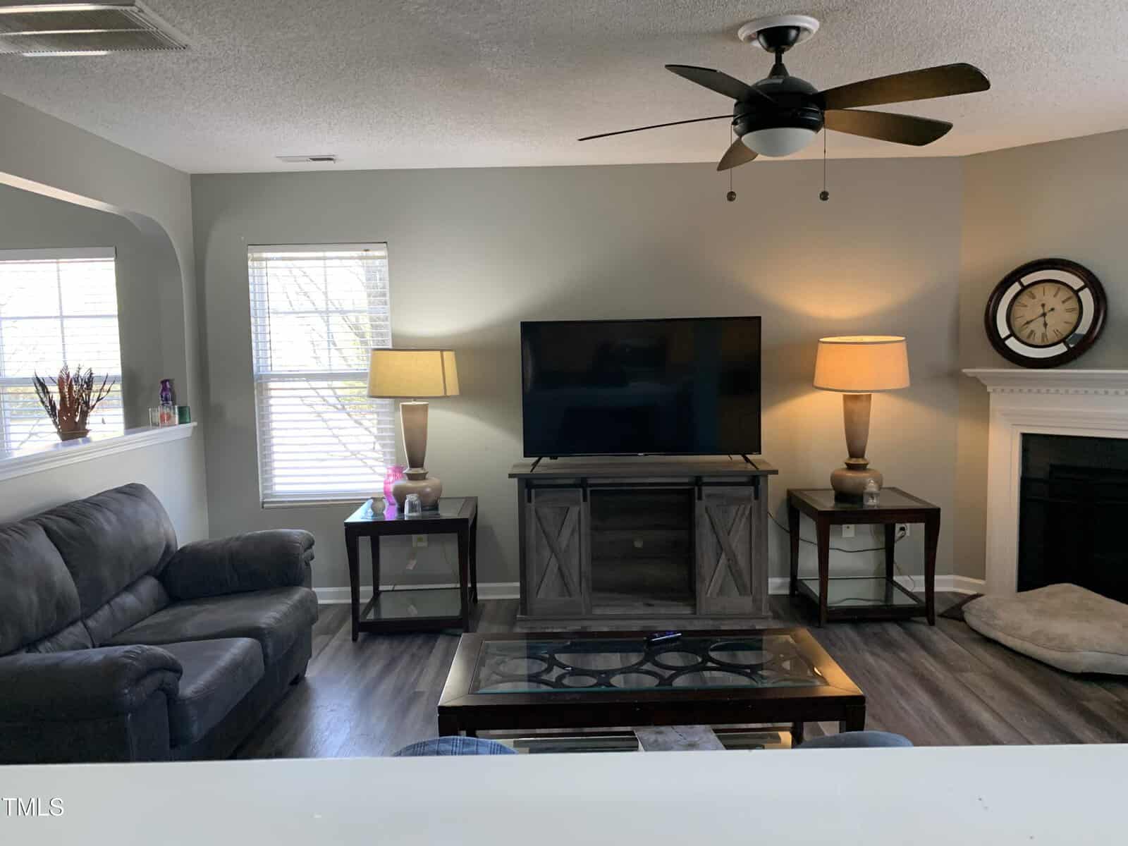 Modern living room with gray walls, featuring a black sofa, glass coffee table, and large flat-screen TV on a wooden stand. Two lamps flank the TV. A ceiling fan above, fireplace on the right, and windows let in natural light.