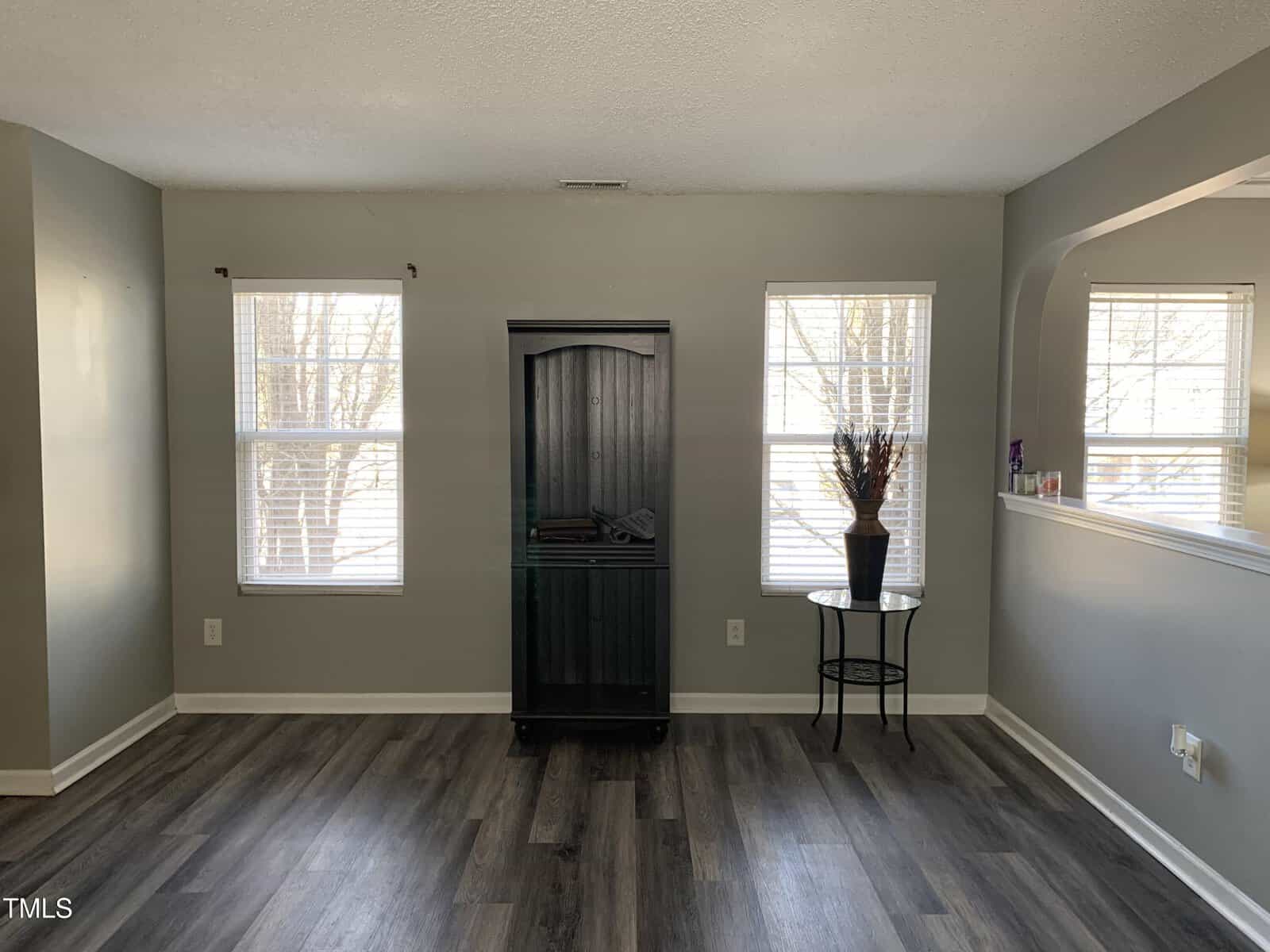 A room with gray walls and wood flooring features two windows, a tall dark cabinet between them, and a small round table on the right. The table holds a vase. An open wall space on the right connects to another room.