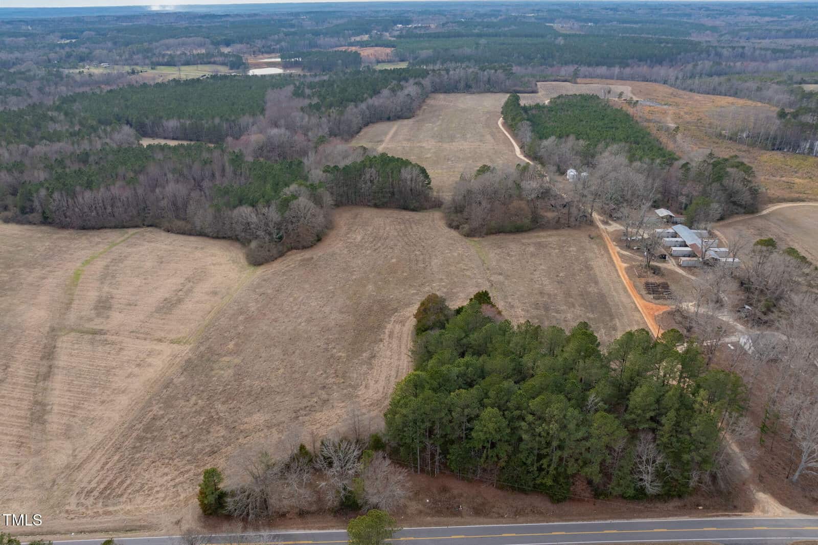 Aerial view of rural landscape showing expansive open fields bordered by patches of trees. A road runs parallel to a section of the fields. On the right, there are a few buildings and a dirt path. Dense forested areas are visible in the distance.