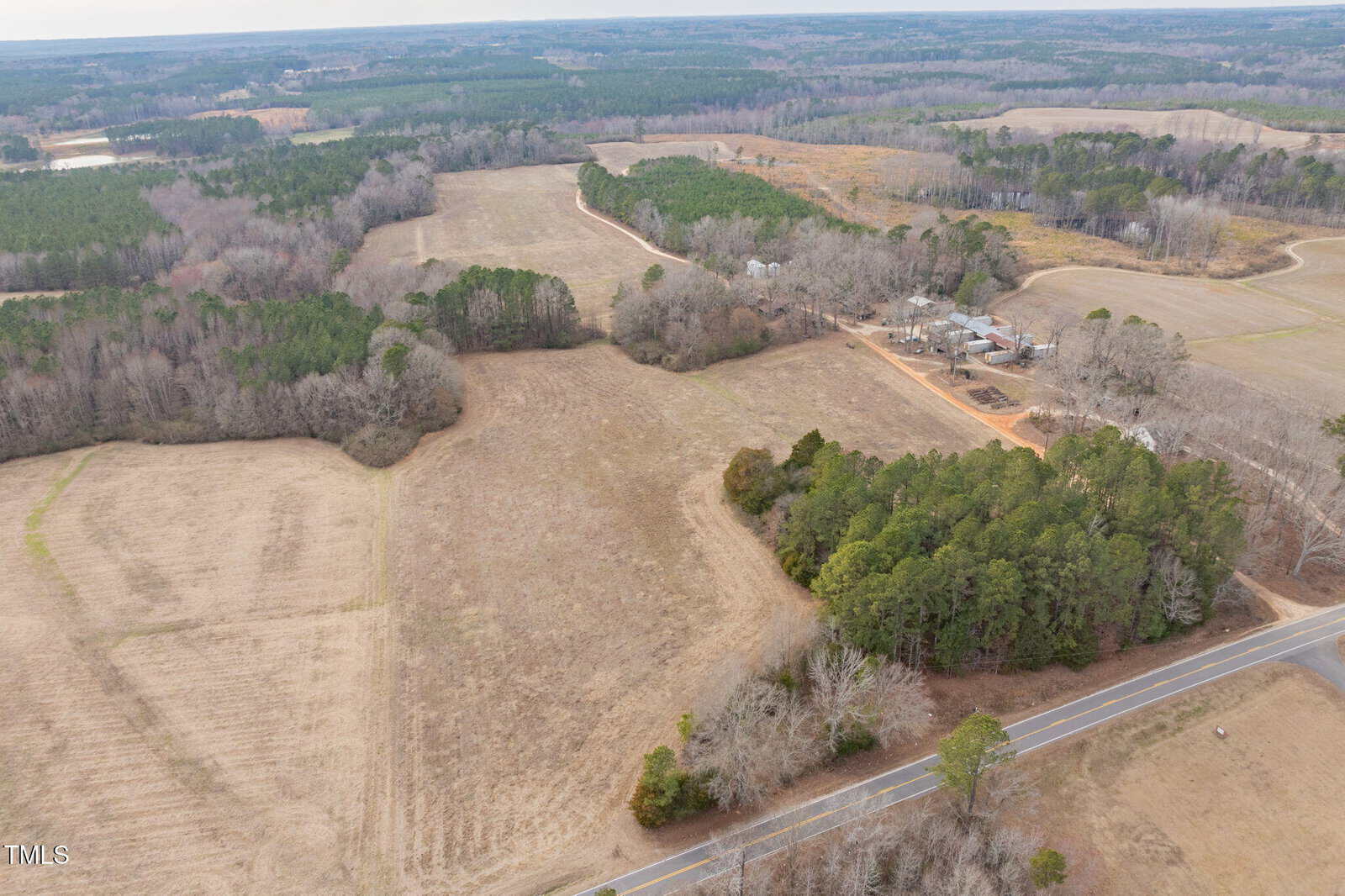 Aerial view of a rural landscape featuring expanses of fields, clusters of trees, and a few scattered houses. A paved road runs diagonally across the bottom of the image, bordered by trees and open land.