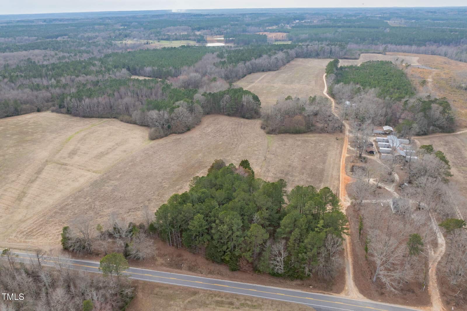 Aerial view of a rural landscape featuring expansive fields, clusters of trees, and a winding dirt road. There are several buildings on the right side. A paved road runs along the bottom of the image, bordered by forested areas.