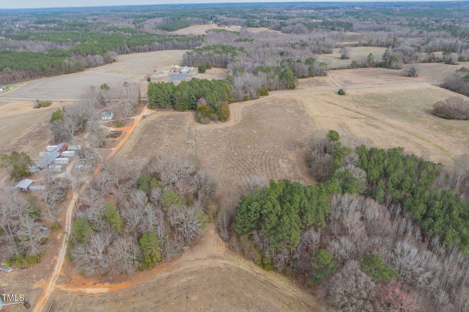 Aerial view of a rural area featuring open fields and patches of forest. Dirt roads can be seen winding through the landscape, and several buildings and farmhouses are visible scattered throughout the area.