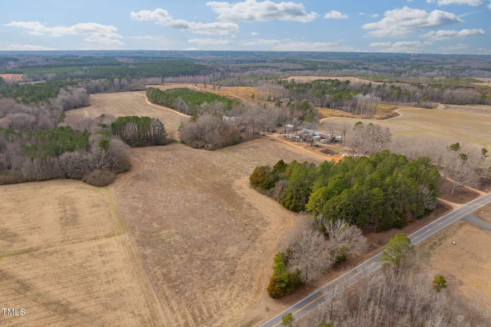 Aerial view of a rural landscape featuring expansive fields, clusters of trees, and a winding road. A small cluster of buildings is visible in the center. The sky is partly cloudy, and the terrain is a mix of brown and green hues.