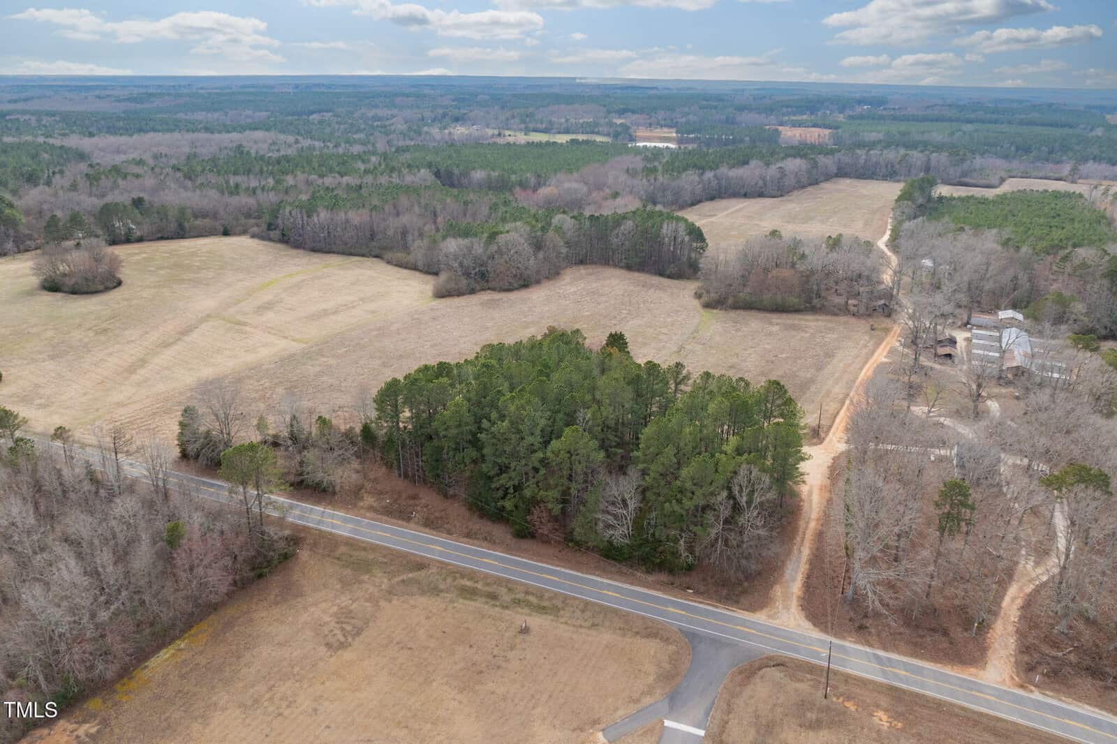 An aerial view of the rural landscape near Henderson reveals open fields, patches of trees, and 2290 Epsom Rocky Ford Road winding through. The horizon features more fields and wooded areas beneath a partly cloudy sky, with a few buildings visible on the right.
