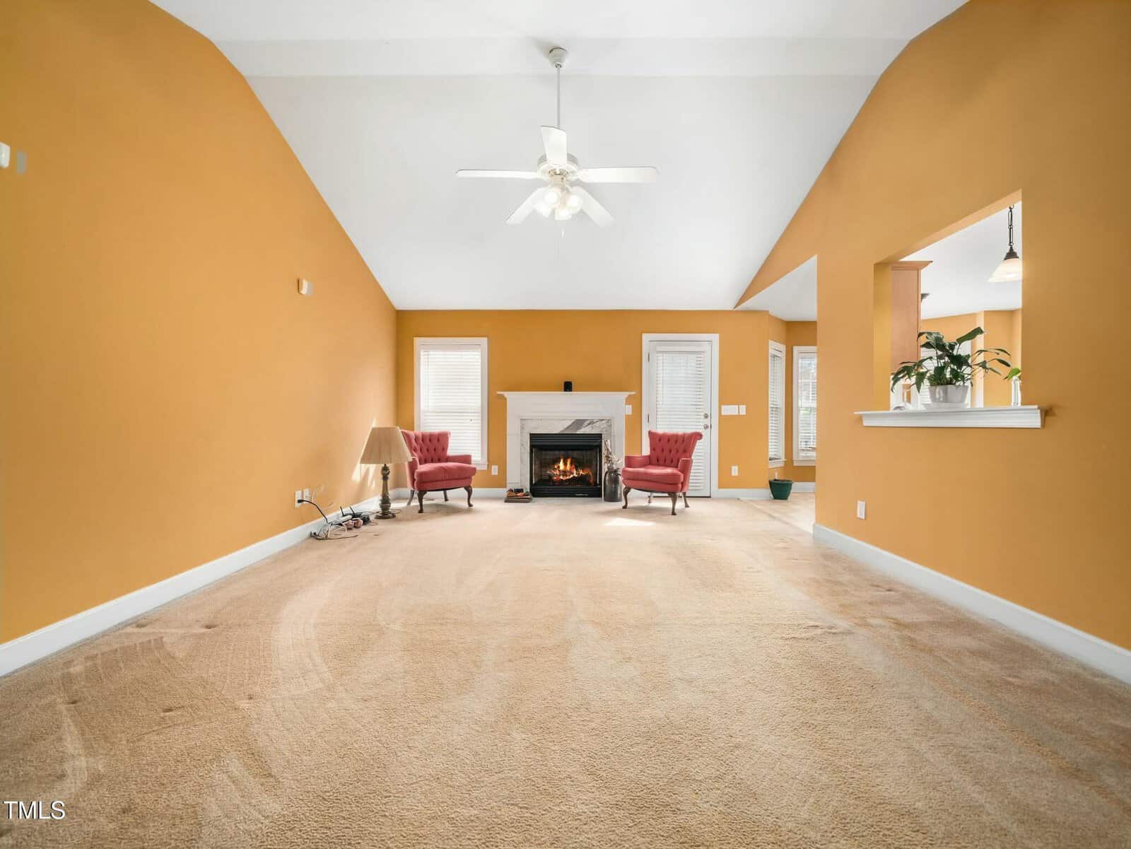 A spacious living room with mustard-colored walls and beige carpet. It features a white fireplace with a lit fire, two red armchairs, and a ceiling fan. The room has a high ceiling and side windows, with a plant on the half wall on the right.