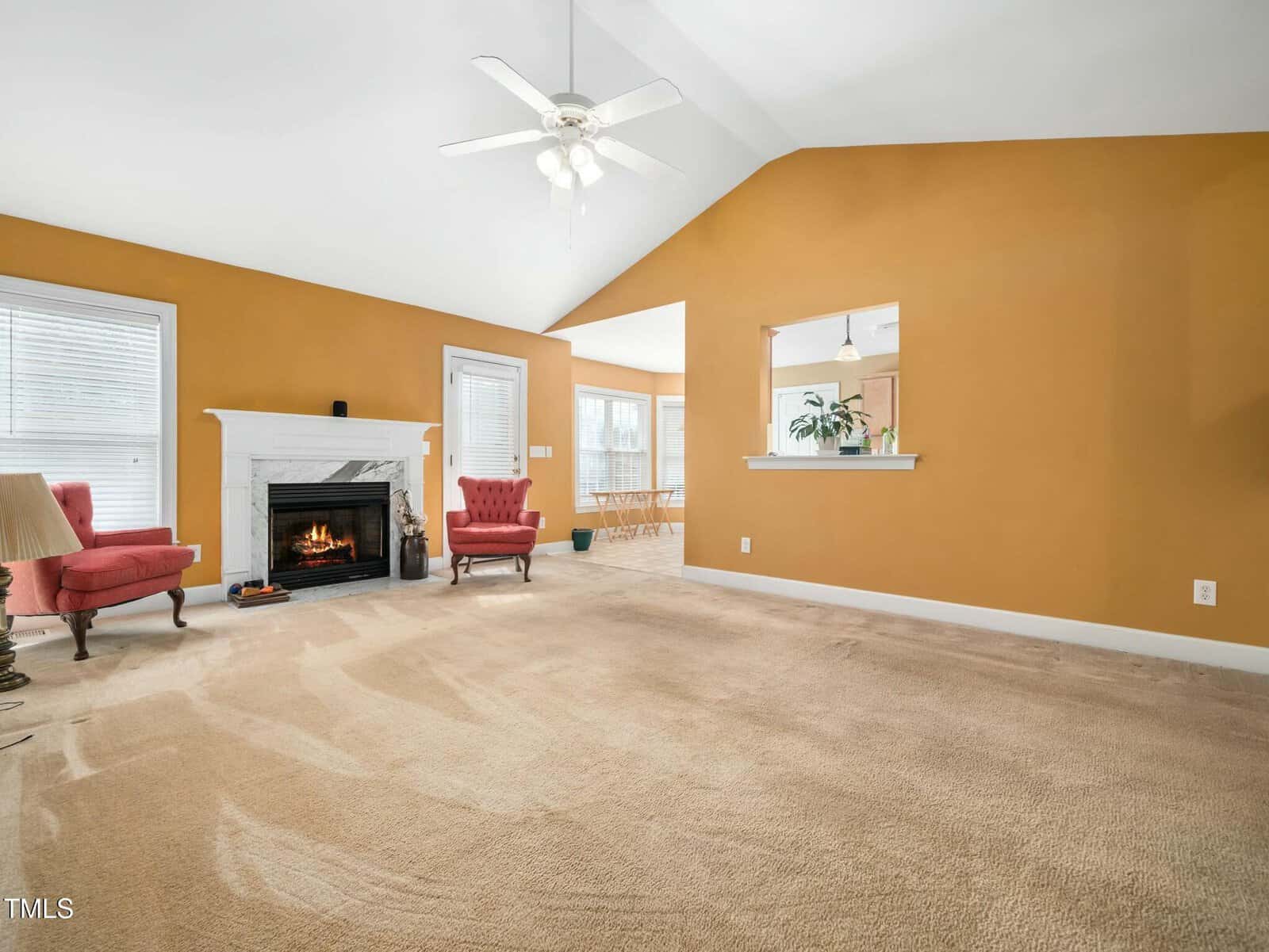 A spacious living room with a vaulted ceiling features mustard-colored walls and beige carpet. It includes a white fireplace with a burning fire, two red armchairs, and a ceiling fan. A large open window reveals an adjacent room with dining furniture.