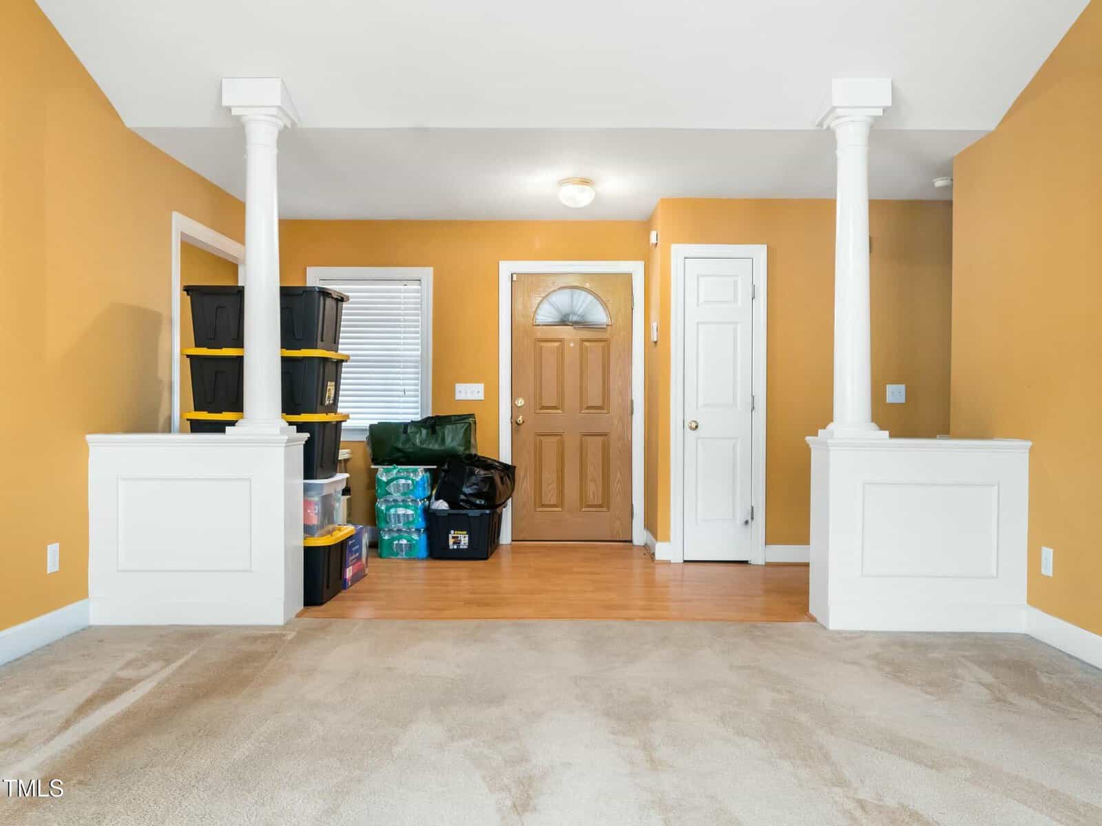 A room with mustard-colored walls features a wooden front door, a white interior door, and two white columns. Carpeted flooring transitions to wood near the doors. Various storage bins and bags are stacked against the wall on the left.