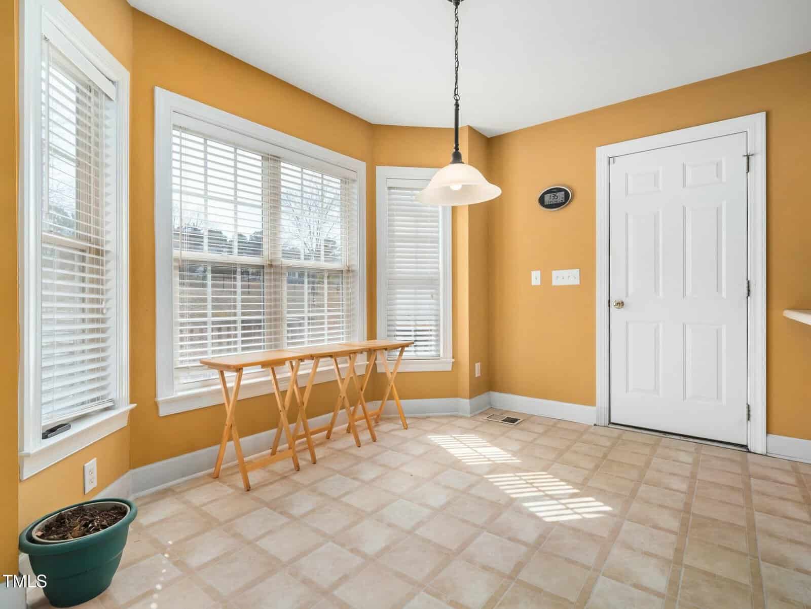 A sunlit room features mustard-colored walls, two windows with white blinds, and tiled flooring. A white door is in the corner, and a wall clock is mounted nearby. Two wooden tray tables are positioned under one window, with a potted plant on the floor.