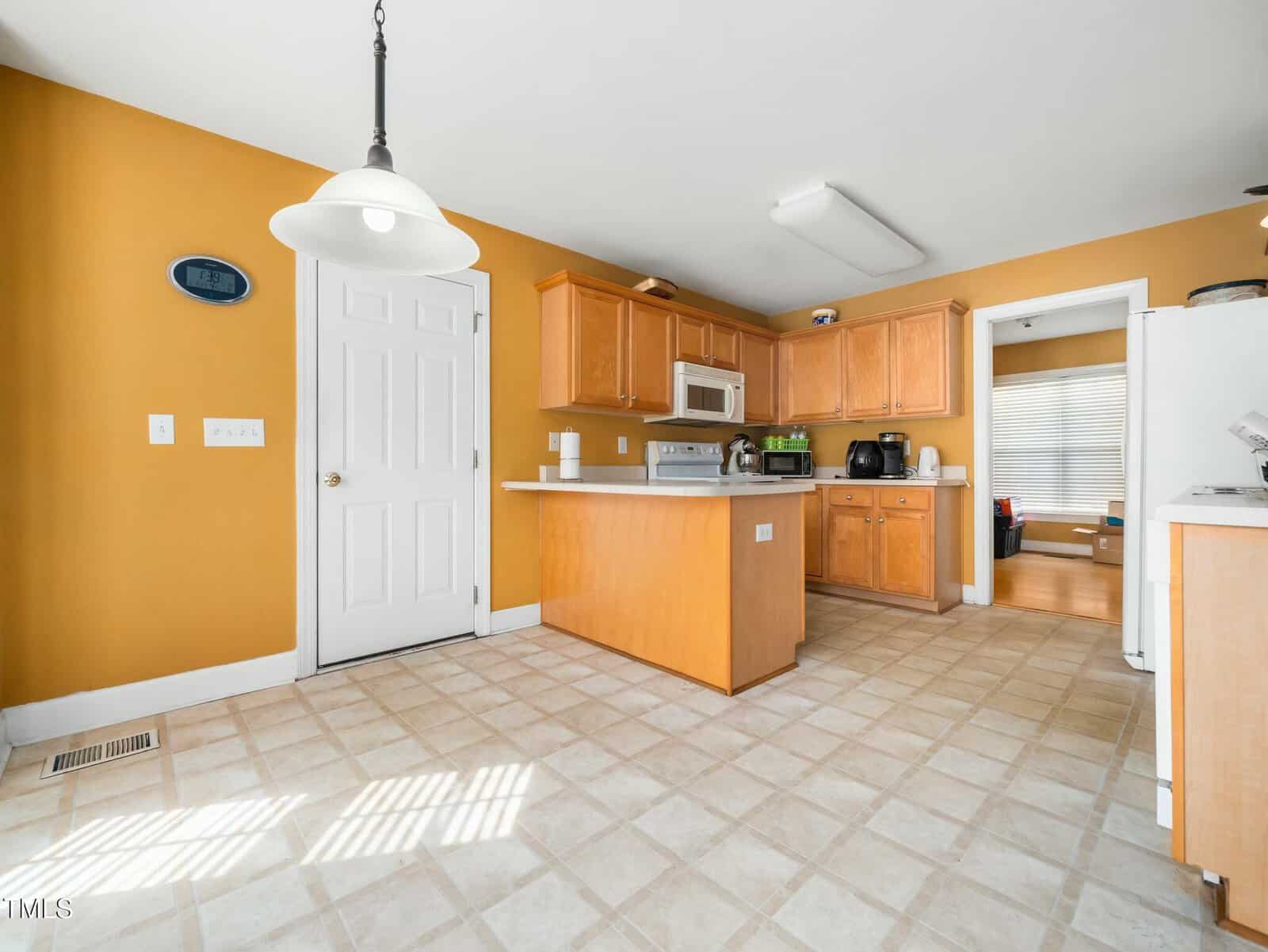 Kitchen with mustard yellow walls, white ceiling, and beige tile flooring. Features include wooden cabinets, a central island, a white door, a hanging light fixture, and various appliances like a microwave and coffee maker. A doorway leads to another room.