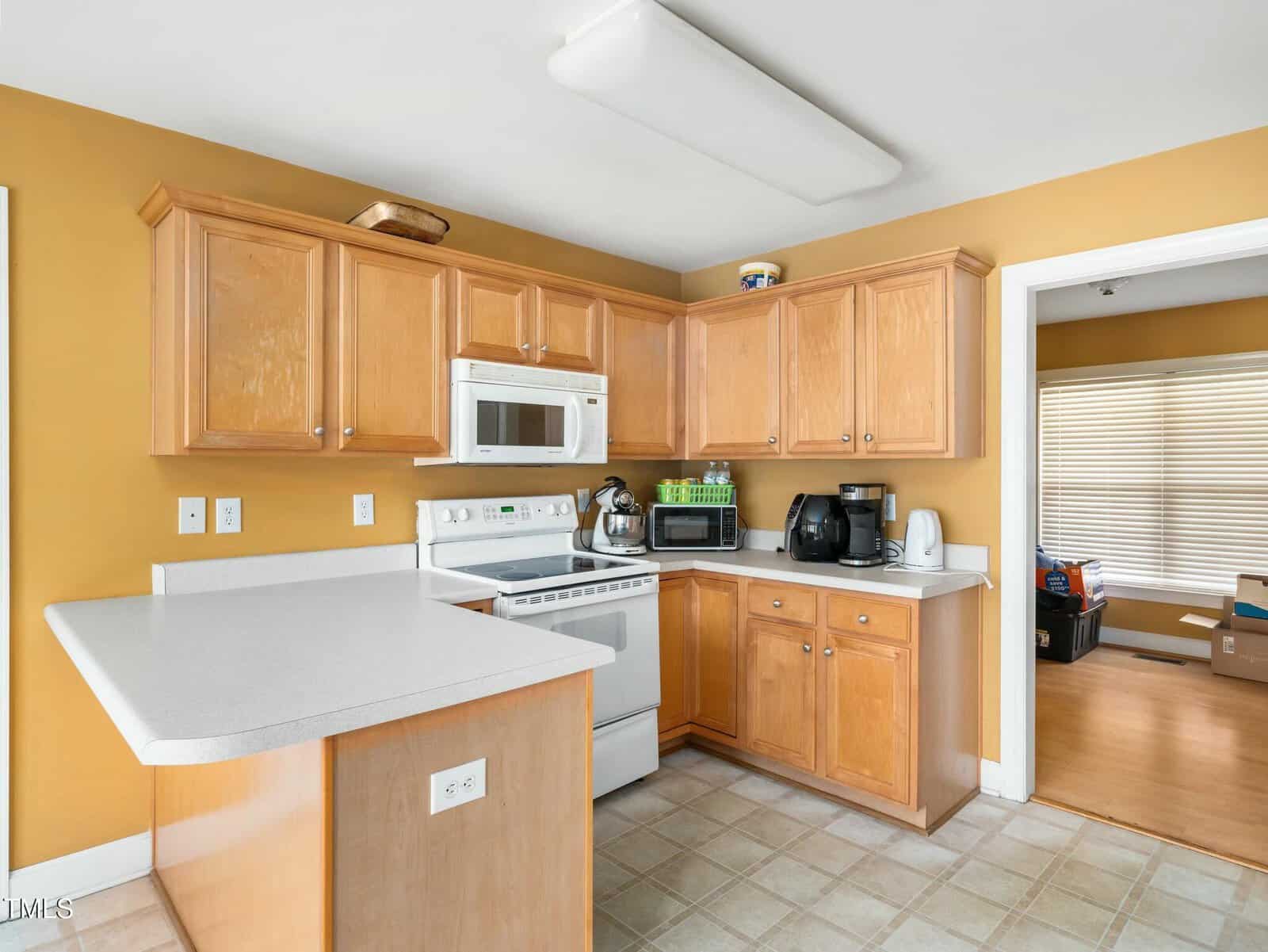 A kitchen with light wood cabinets, white countertops, and tiled flooring. Appliances include a white microwave, stove, toaster, and coffee maker. The walls are painted yellow. An adjacent room with unpacked cardboard boxes is visible through an open doorway.