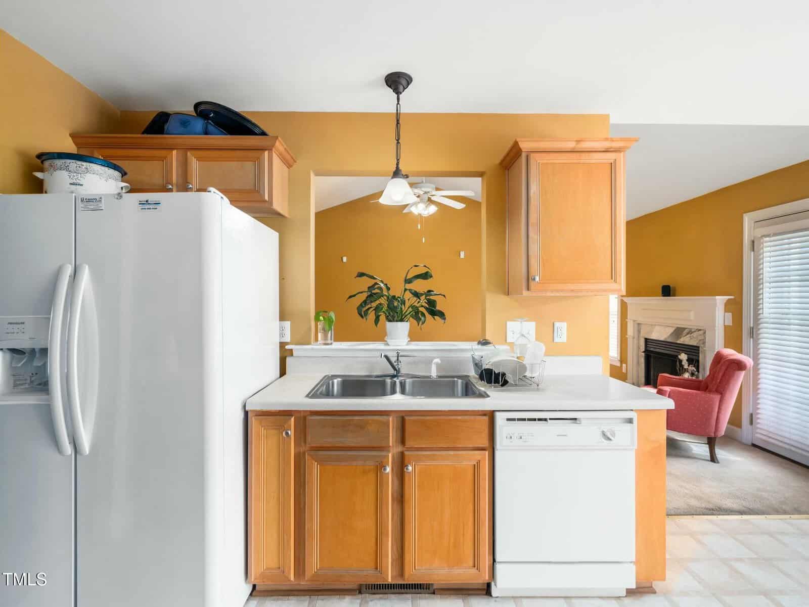 Kitchen with wooden cabinets, a white refrigerator, and a dishwasher beneath a double sink. A potted plant is near the sink. The walls are a mustard yellow. A red armchair is near a fireplace in the adjoining room. Ceiling fan with a light above.