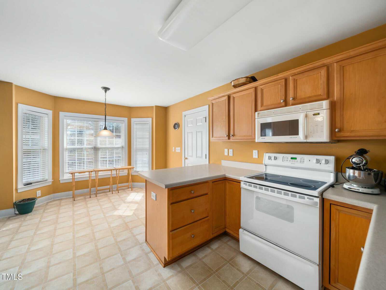 A spacious kitchen with light wood cabinets and beige countertops. It features a white stove and microwave. A mixer is on the counter. A dining area with a large window and a table with chairs is in the background. The walls are painted yellow.