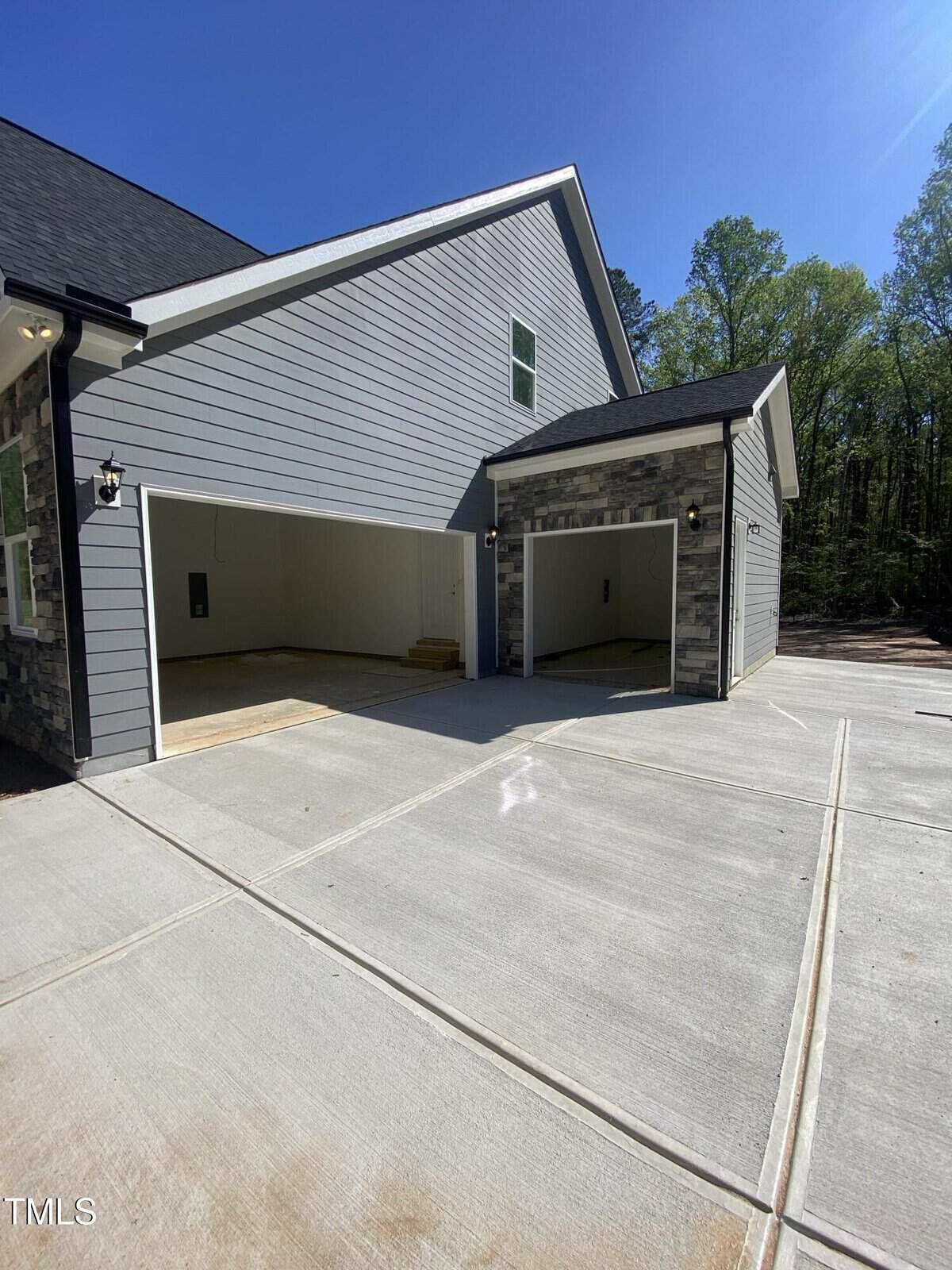A newly constructed house with two open garage doors, gray siding, stone accents, and a large concrete driveway. Trees are visible in the background under a clear blue sky.