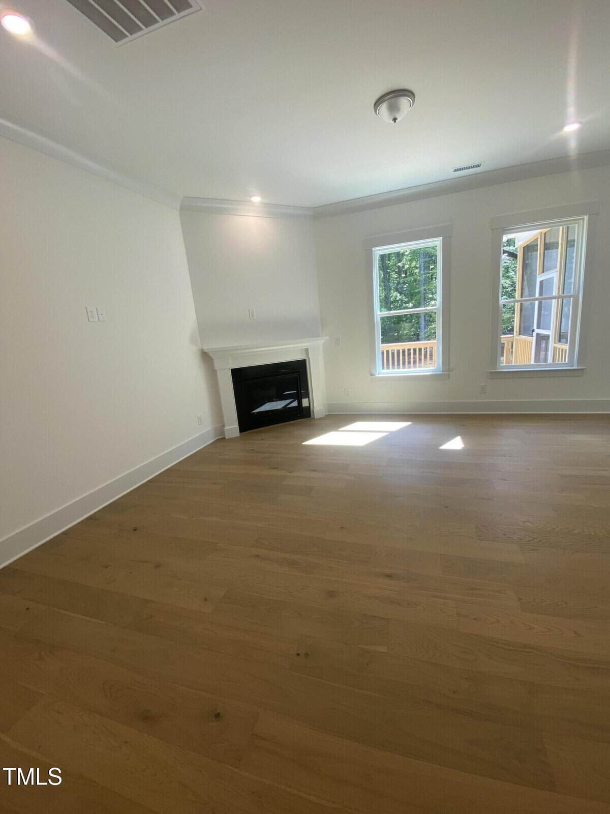 A bright, empty room with light wood floors, white walls, a corner fireplace, and two tall windows letting in natural light. A small portion of a deck is visible outside the windows.