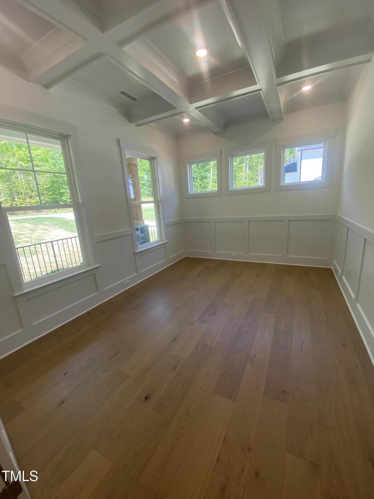 Empty room with light wood flooring, white walls with paneling, a coffered ceiling, and several windows letting in natural light. Trees and grass are visible outside the windows.