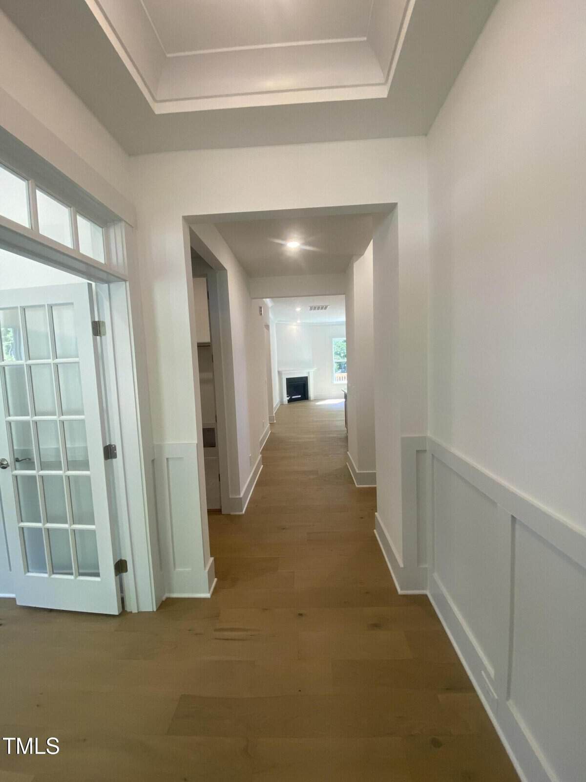 A hallway with light wood floors, white walls, wainscoting, a French door on the left, and a distant view of a fireplace and windows at the far end. Ceiling lights are on, and natural light enters from the windows.