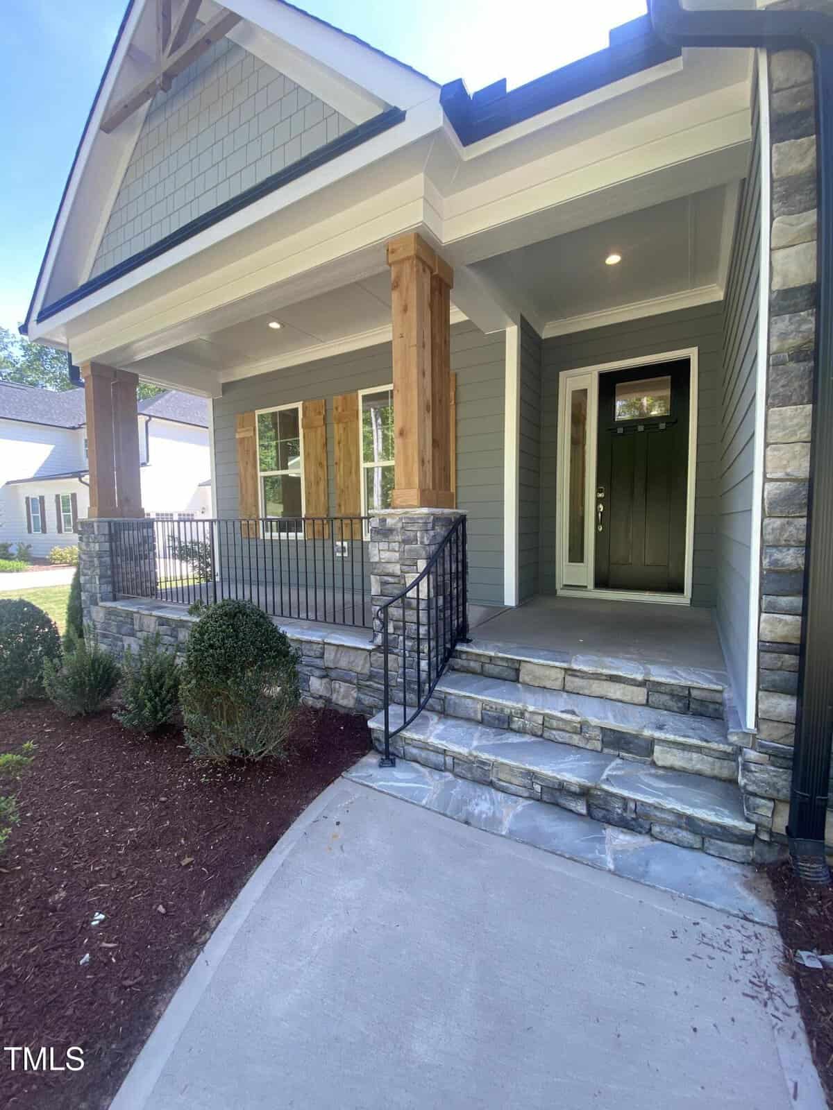A single-story house with a covered front porch featuring wooden columns, stone steps, and a black metal railing. The exterior has gray siding, stone accents, and landscaped bushes along the walkway.