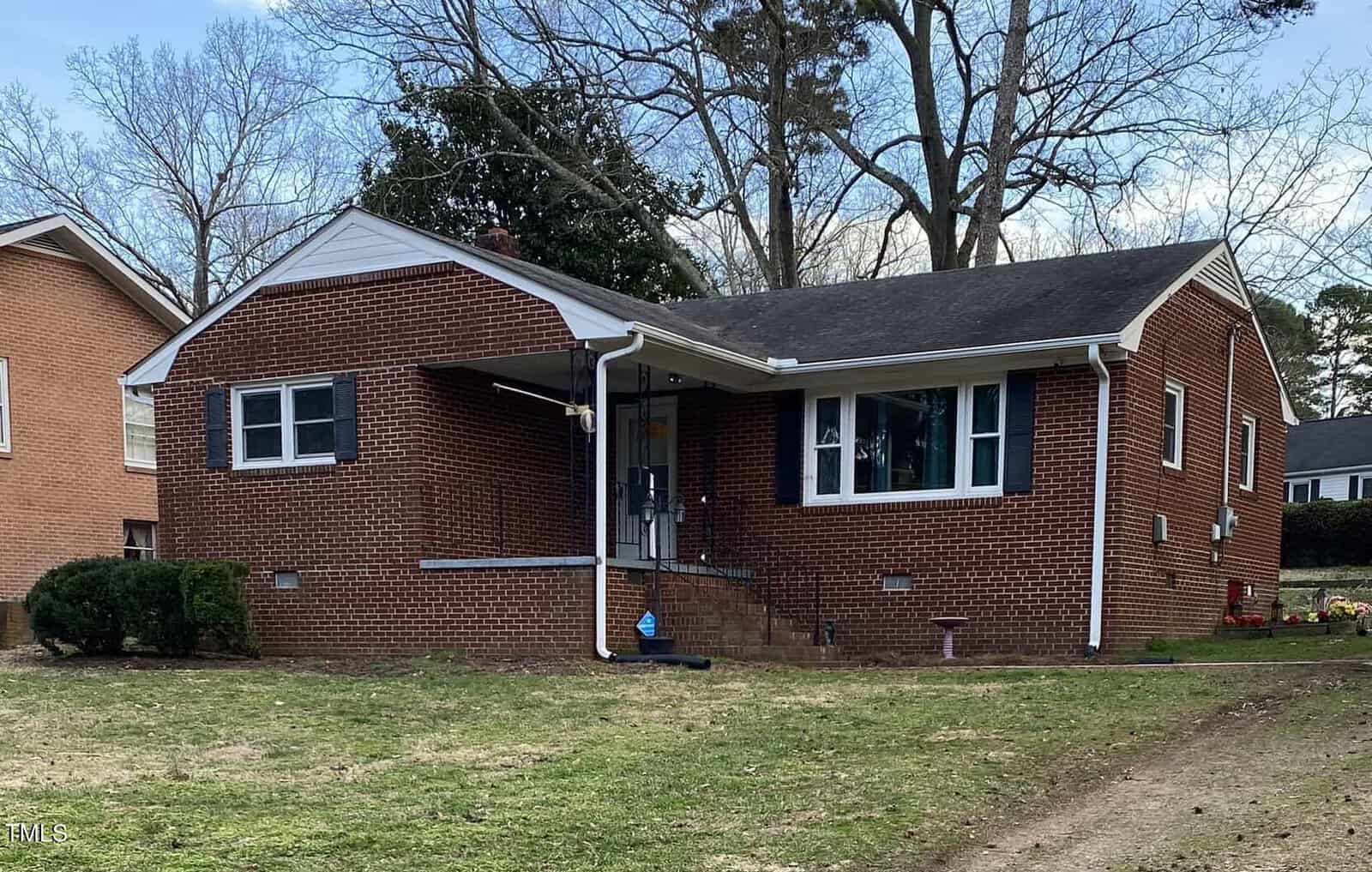 A red brick single-story house with a small front porch and steps leading up to it. The house has a sloped roof and several windows. Bare trees are visible in the background, and the front yard is mostly grass with some bushes near the house.