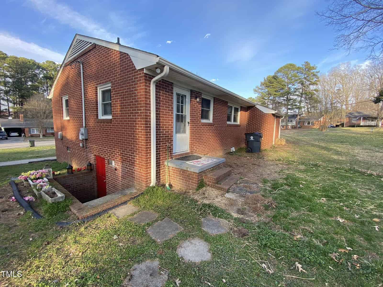 Single-story brick house with a side entrance door and small concrete steps. A set of steps leads down to an under-house door. The yard is grassy with some bare patches, and there are flowering plants near the steps. Trees and houses are in the background.
