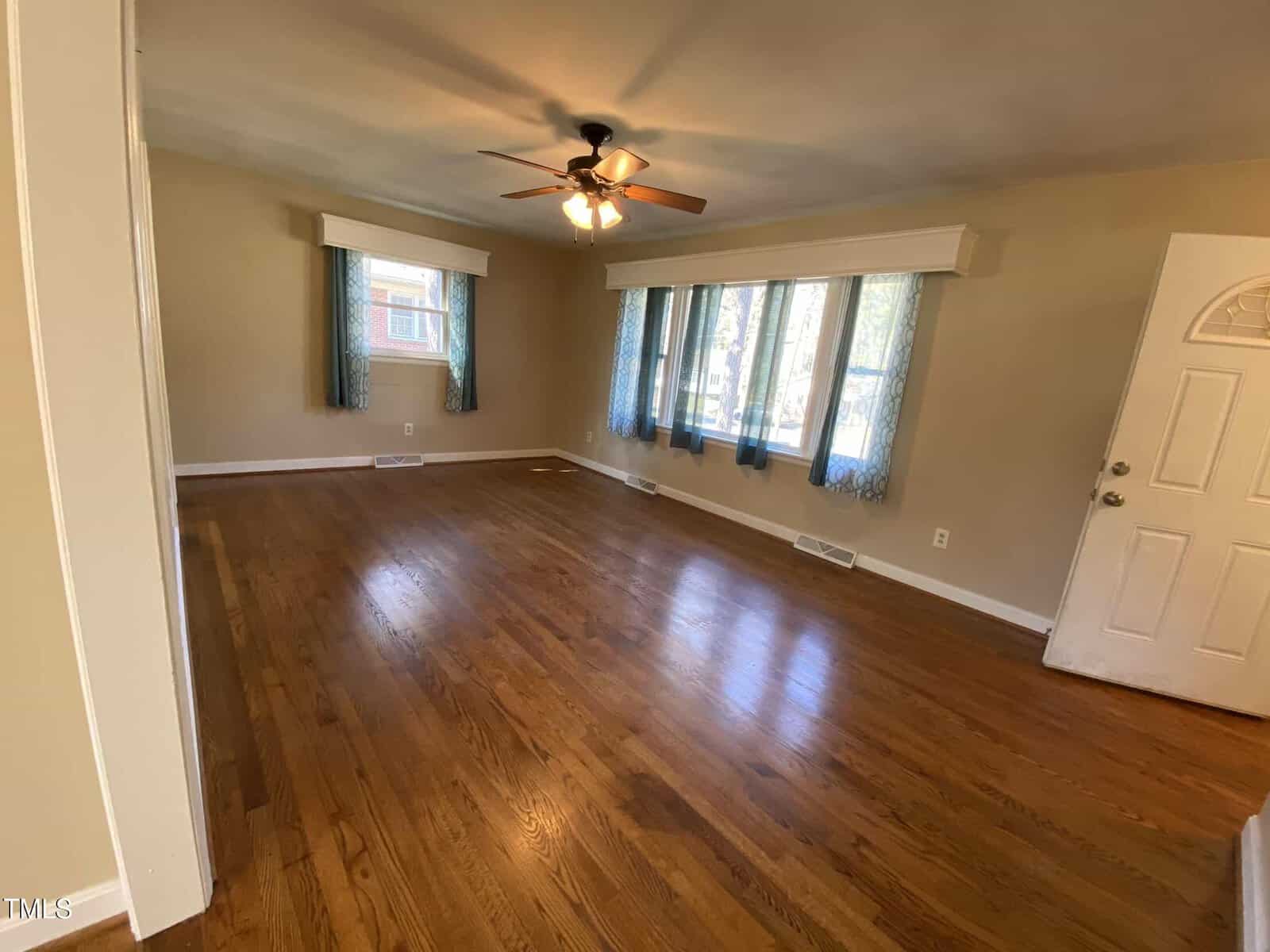 A spacious living room with polished hardwood floors and beige walls. A ceiling fan with lights is centered on the ceiling. Three windows with blue curtains let in natural light, and a white door is visible on the right.