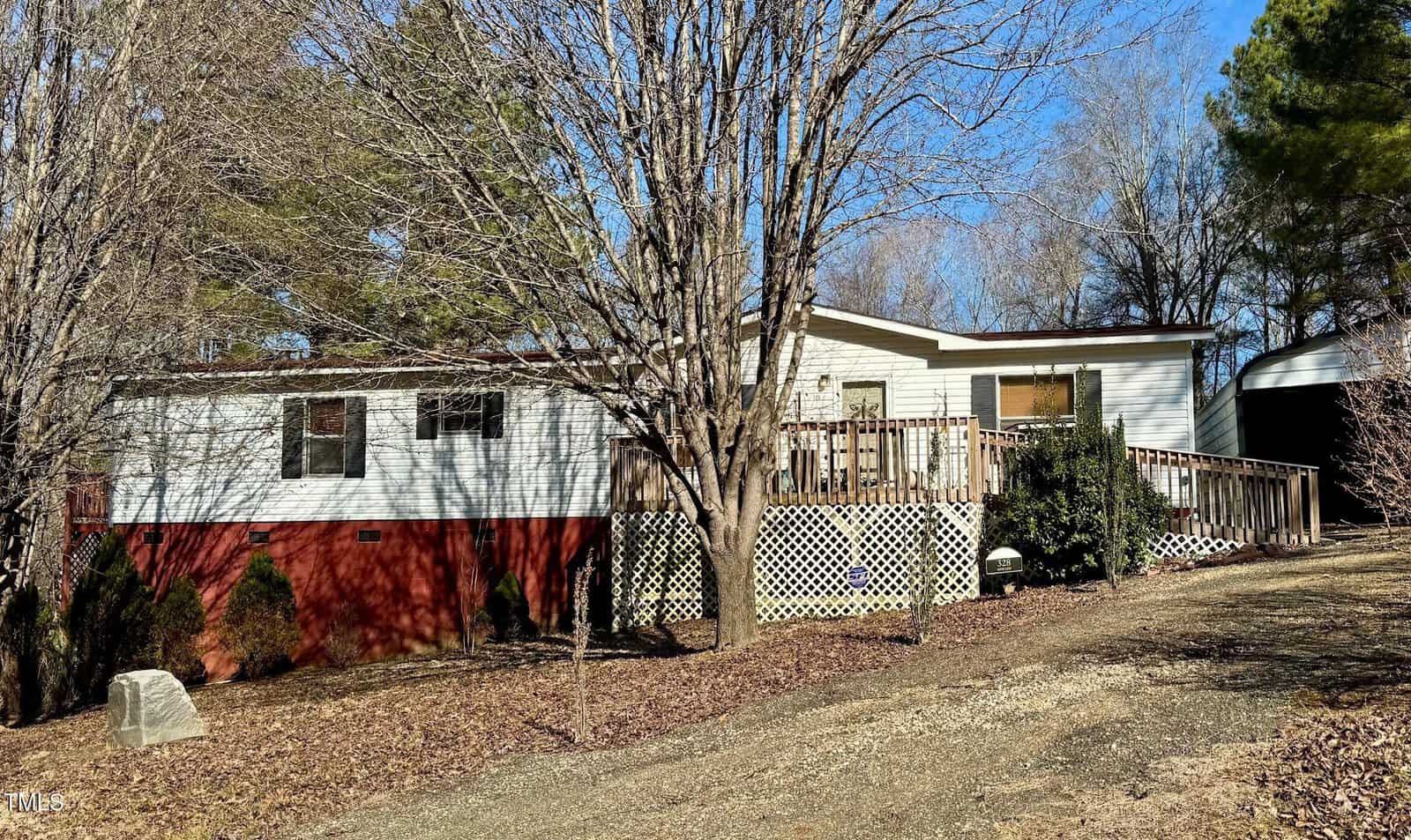 A single-story mobile home with white siding and a red foundation is surrounded by bare trees. There is a wooden deck attached to the front of the house. The yard is covered in dry leaves and a sloped gravel path leads to the entrance.