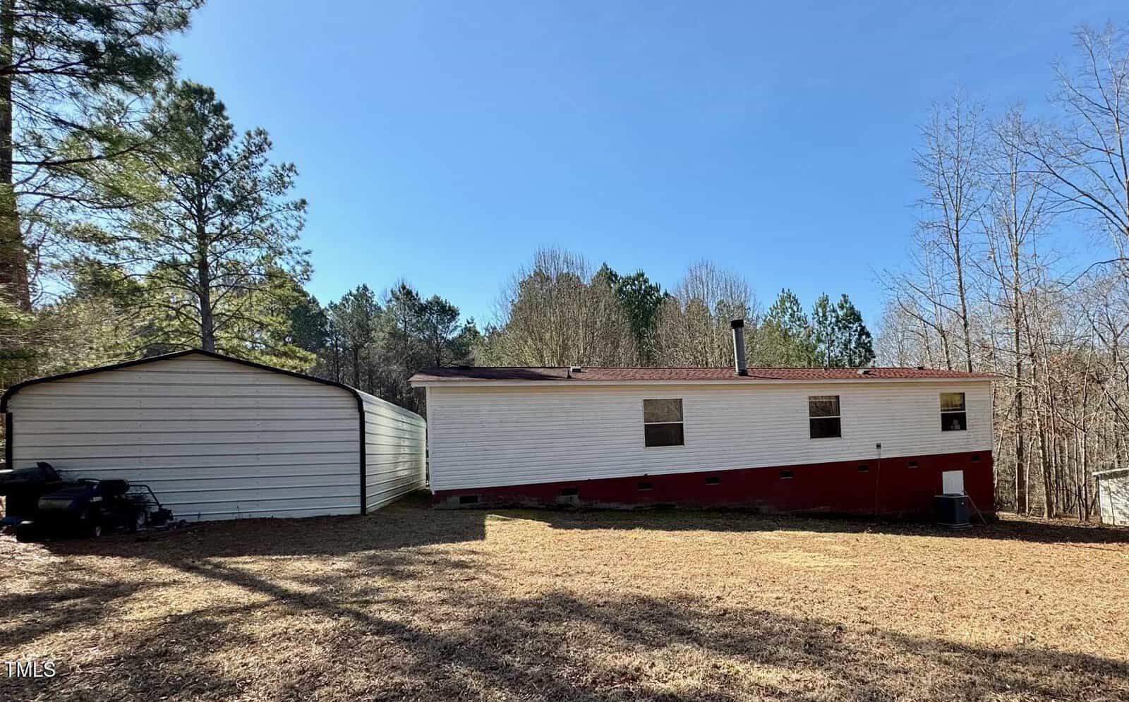 A single-story mobile home with a white upper section and red lower section sits on a grassy lot. A detached white garage is on the left. The area is surrounded by trees under a clear blue sky.