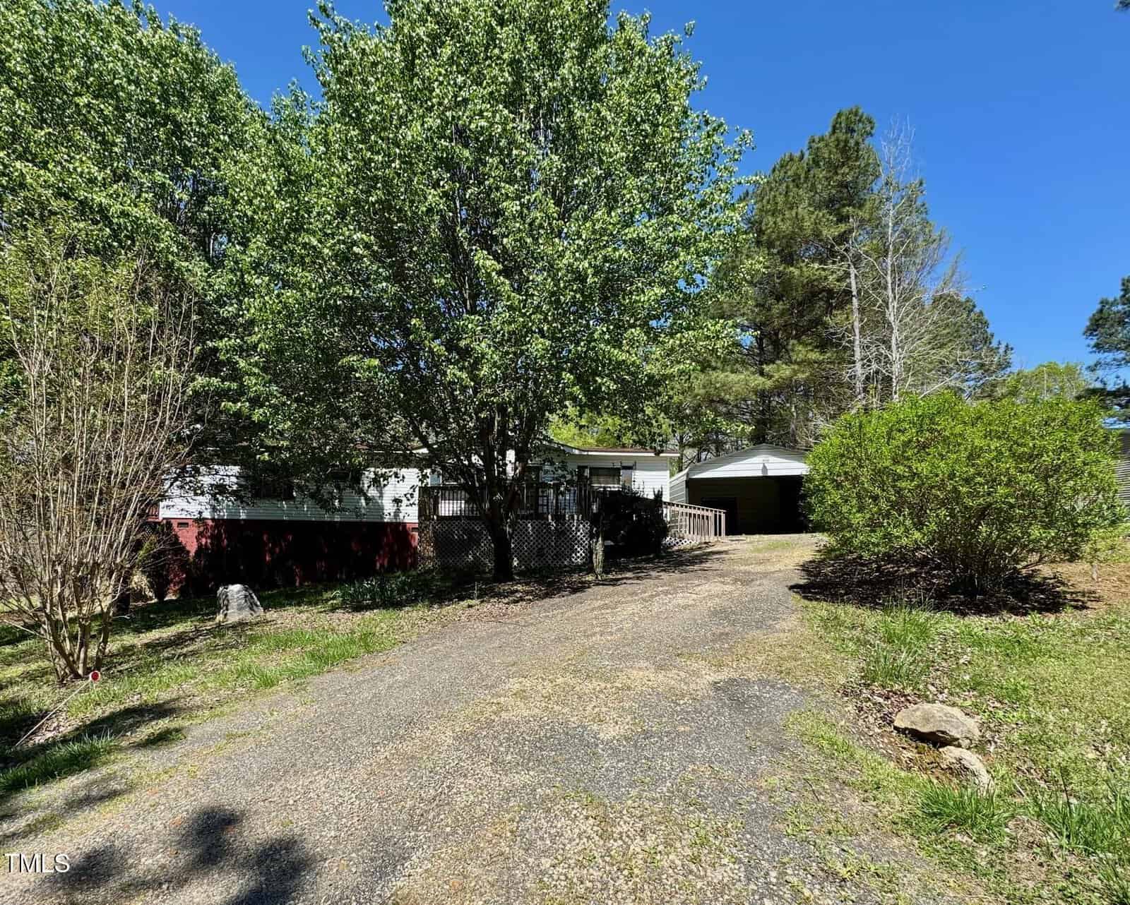 A gravel driveway leads to a small white house with a carport, partially obscured by large green trees and bushes. The sky is clear and blue, and the surrounding yard is grassy with patches of sunlight and shadow.