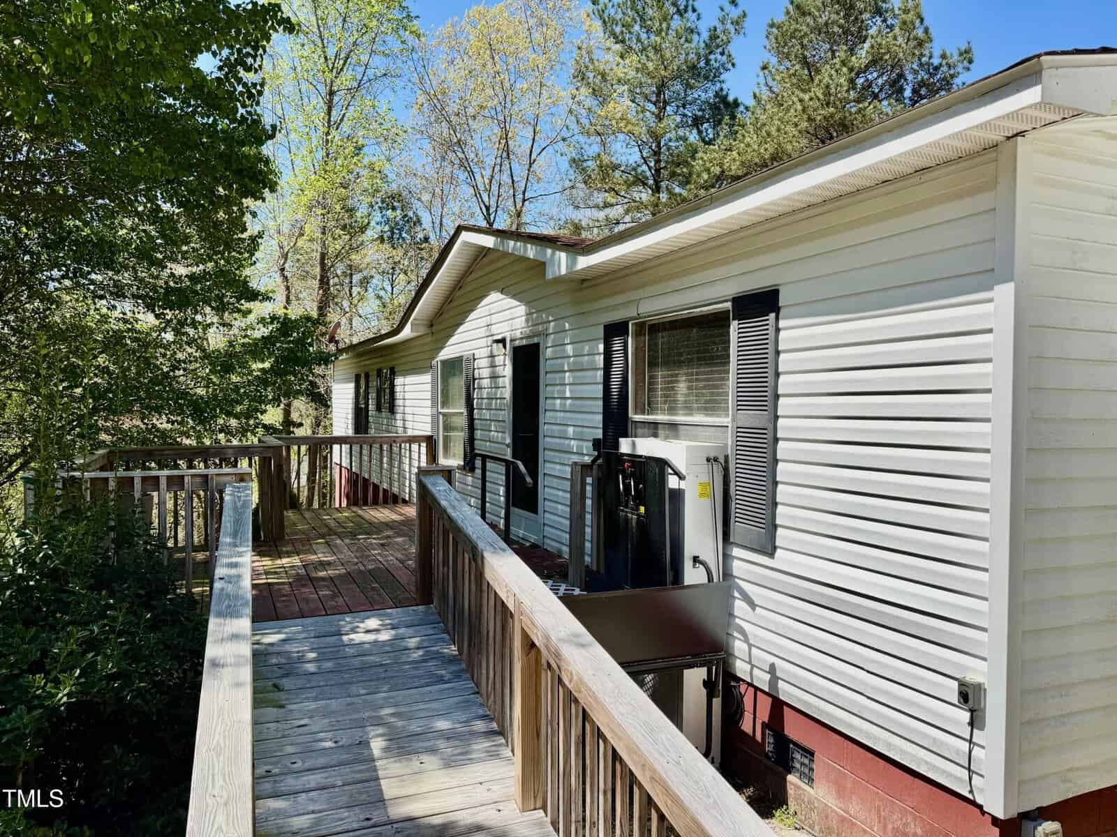 A white mobile home with a sloped wooden ramp leading to the entrance. The exterior features small windows with shutters and is surrounded by tall trees under a clear blue sky.