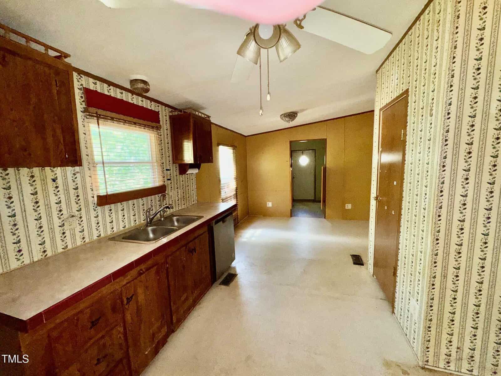 A kitchen with wooden cabinets, a white countertop with a sink, and patterned wallpaper. The ceiling has a fan and light fixture. There are windows on the left, and a corridor with doorways at the far end.