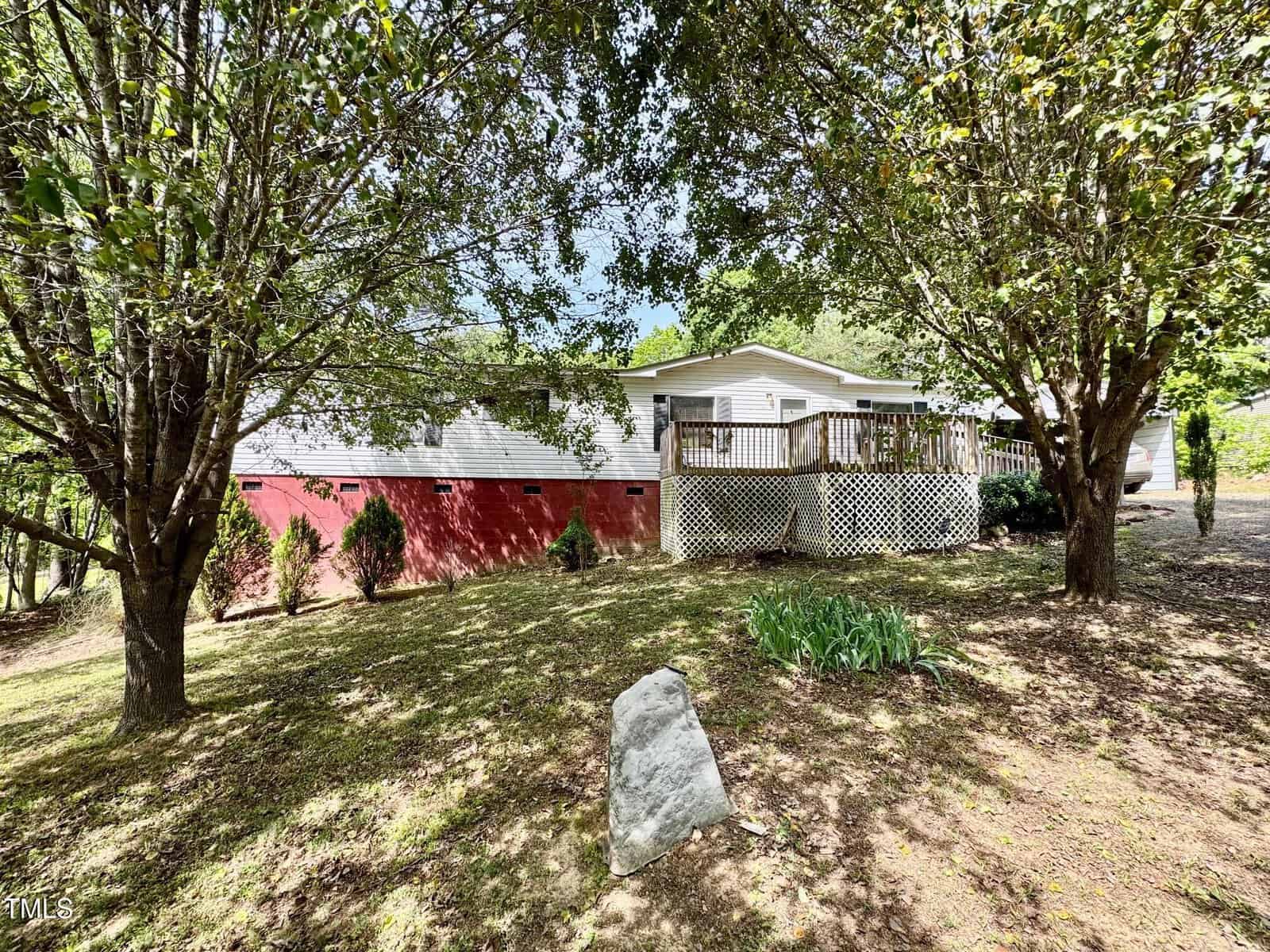 A single-story house with white siding and a red brick foundation sits behind two large leafy trees. The house has a wooden deck with railings and steps. The front yard features grass, shrubs, and a large rock.