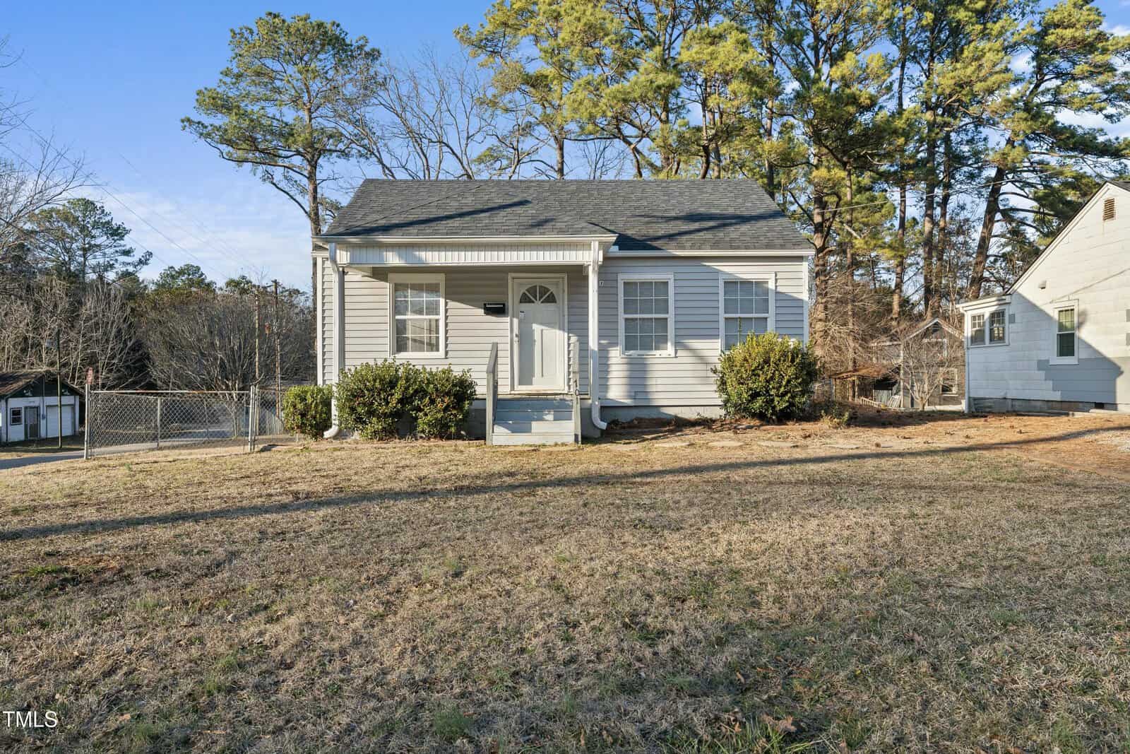 A small, light gray house with white trim and a covered front porch sits on a grassy, slightly sloped yard with bushes and trees in the background under a clear blue sky. A white house is visible to the right.