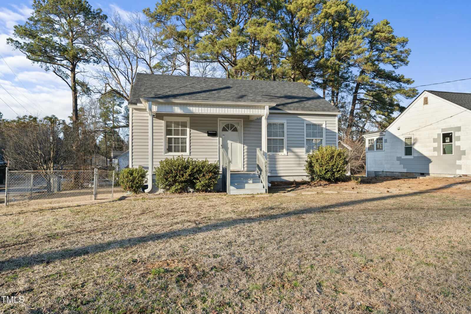 Single-story gray house with white trim, a small front porch with steps, bushes along the front, and a fenced yard. There are trees and a neighboring house in the background.