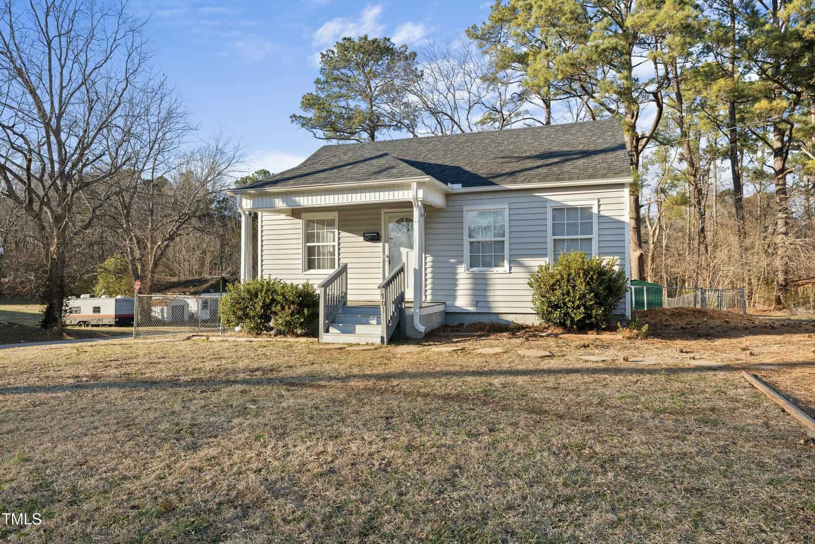 A small, single-story house with gray siding and a covered front porch, set on a grassy lawn with a few bushes and surrounded by trees under a clear sky.