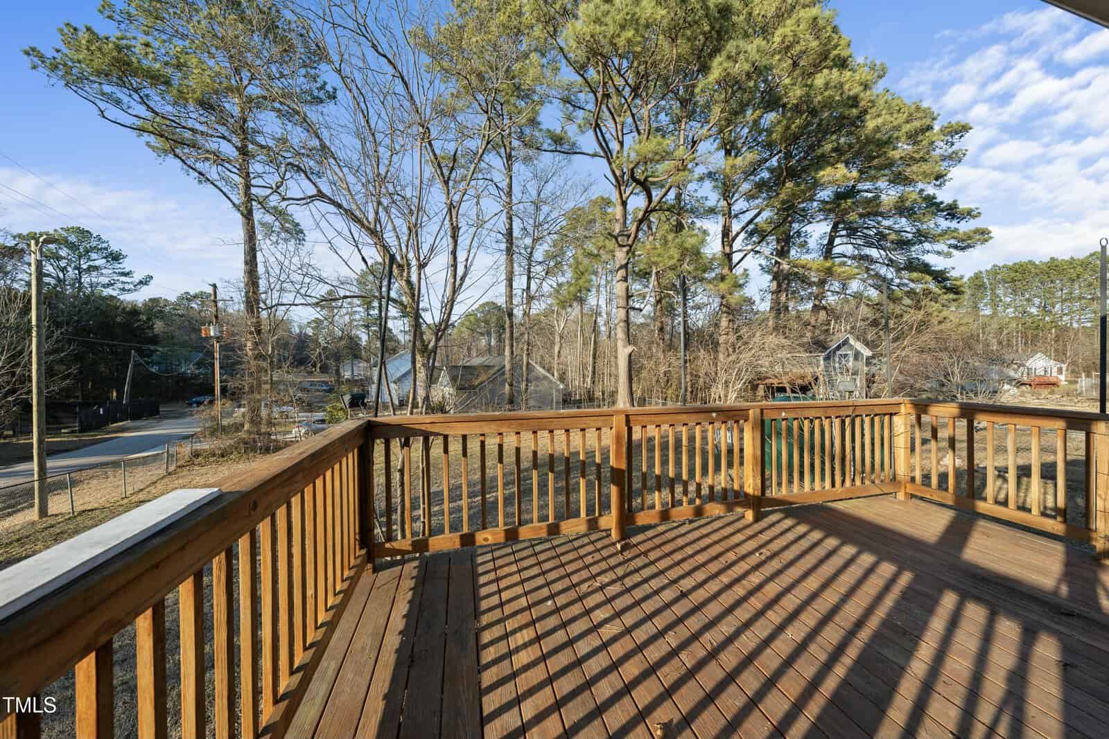 A wooden deck with a railing overlooks a yard with trees and a residential street. Several houses are visible in the background under a partly cloudy sky. Shadows from the railing fall across the deck.