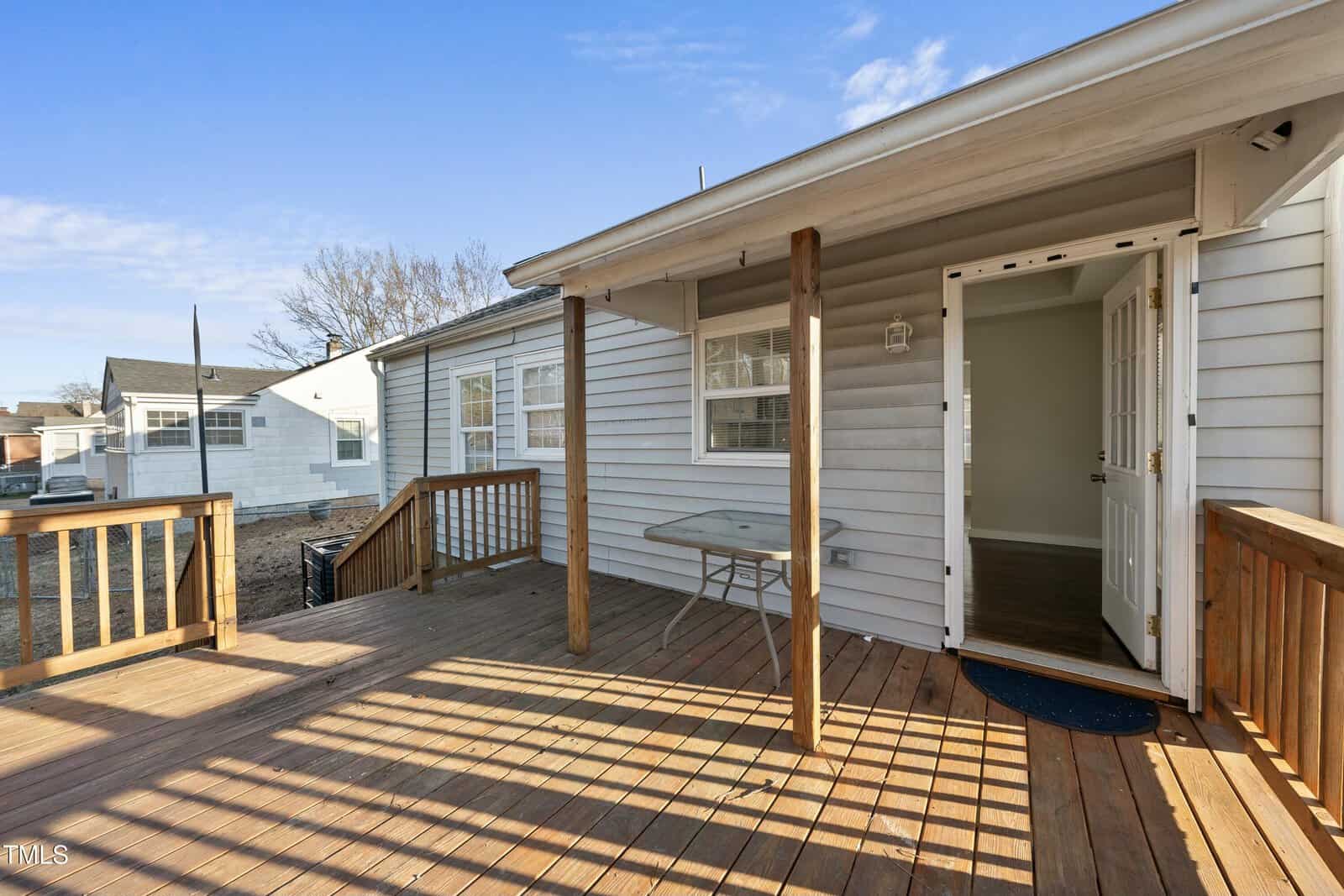 A wooden deck attached to a white house with siding, featuring a small table, wooden railings, and an open door leading inside. Other houses and a clear blue sky are visible in the background.