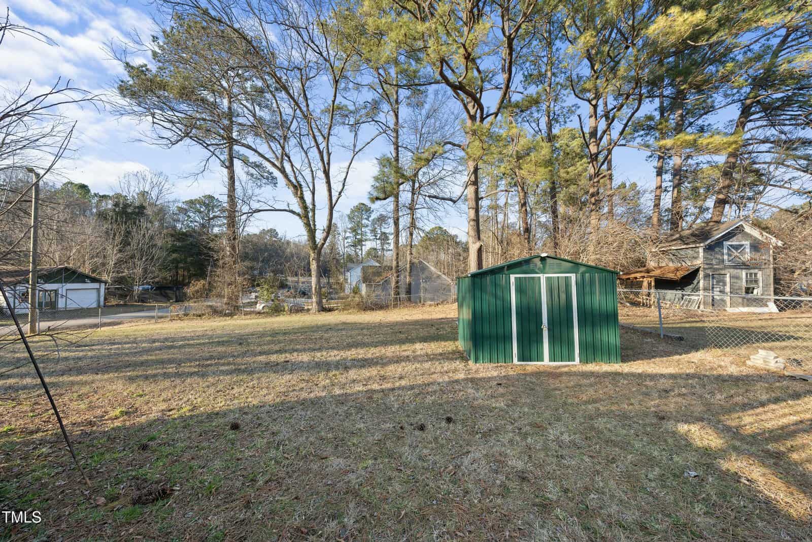 A green metal shed sits in a large, grassy backyard bordered by a chain-link fence, with tall trees and neighboring houses visible in the background under a clear sky.